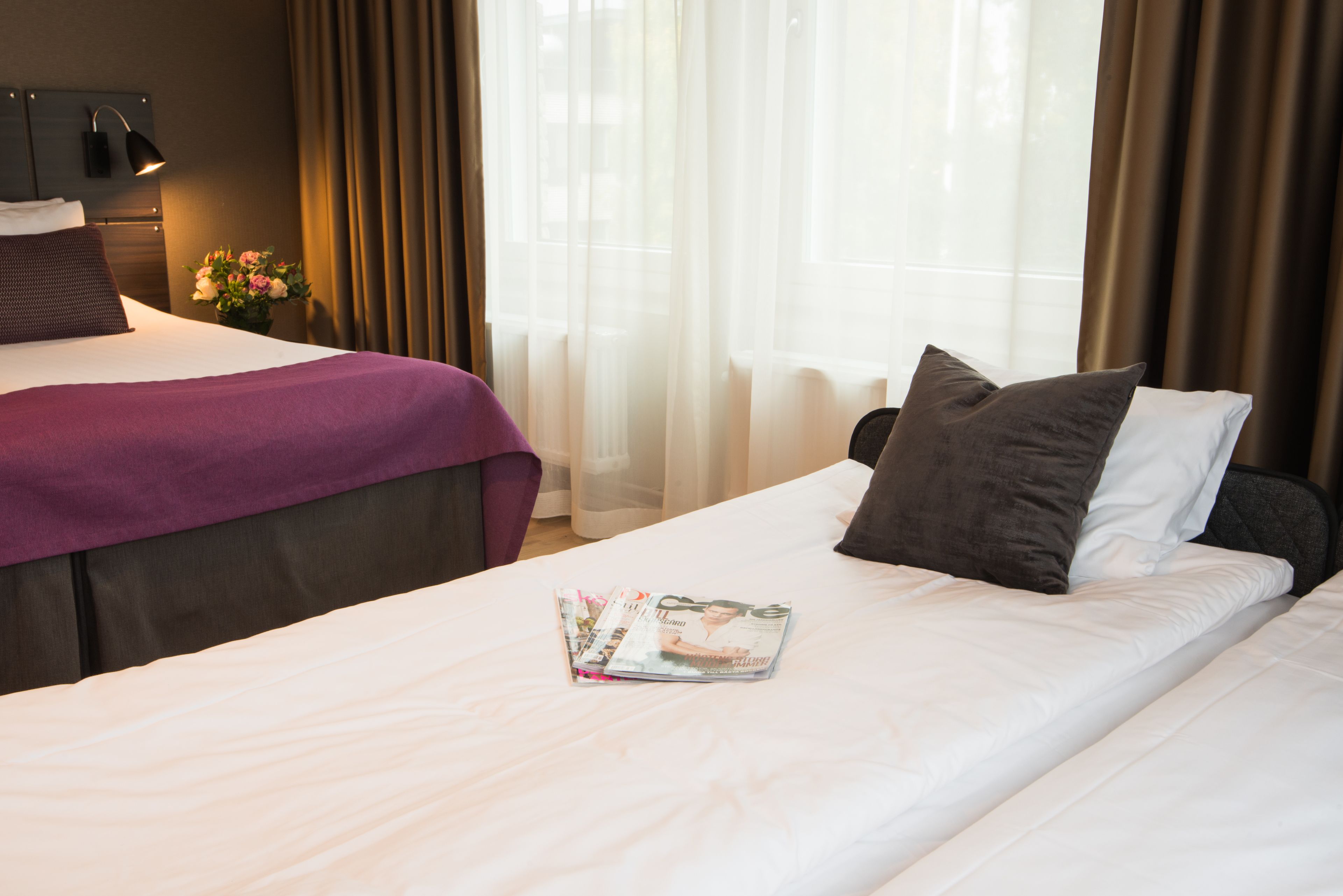 Una habitación de hotel con una cama, una mesita de noche con lámpara, una ventana con cortinas y un jarrón con flores.