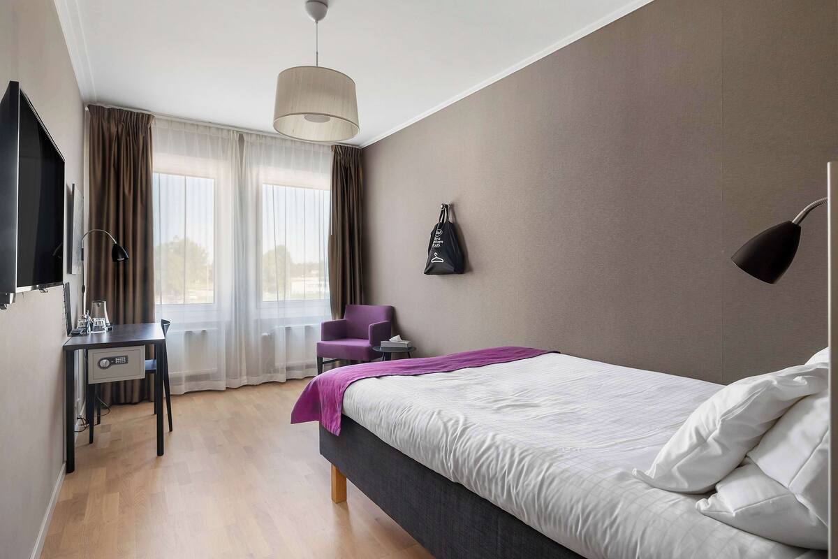 A hotel room with a bed, a desk, a chair, a TV, and a window with curtains.
