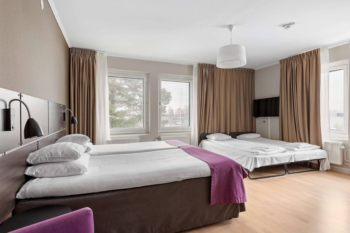 A hotel room with two single beds, a large window, a flat-screen TV, and a ceiling-mounted light fixture.