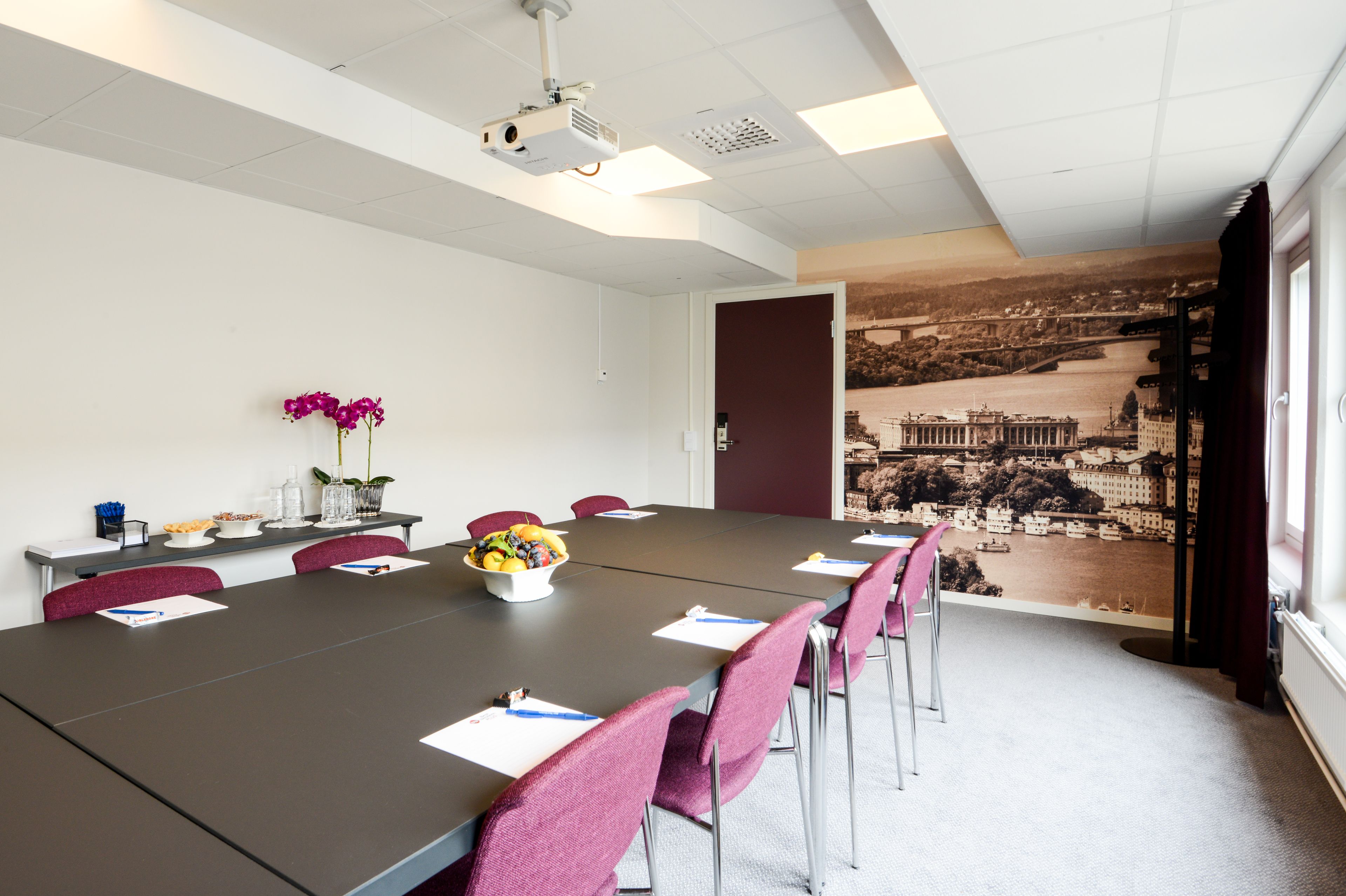 A conference room with a long table, chairs, a projector, and a mural of a cityscape on the wall.