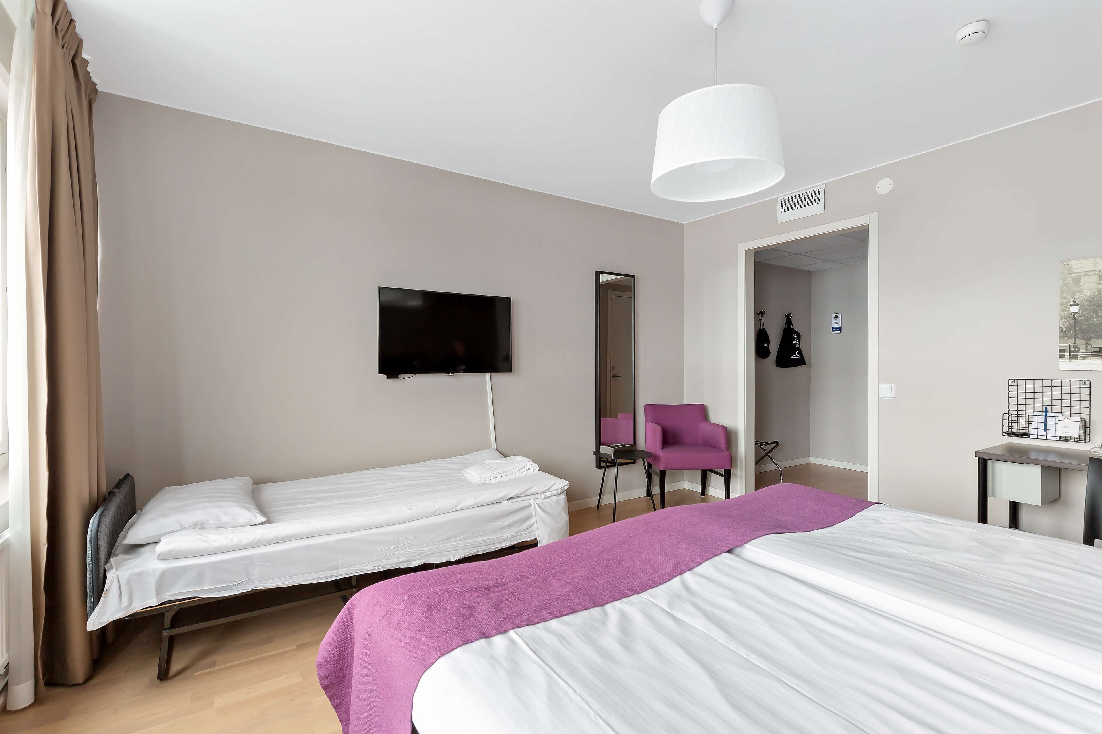 Habitación de hotel con dos camas individuales, televisión, espejo, una silla y escritorio con lámpara.