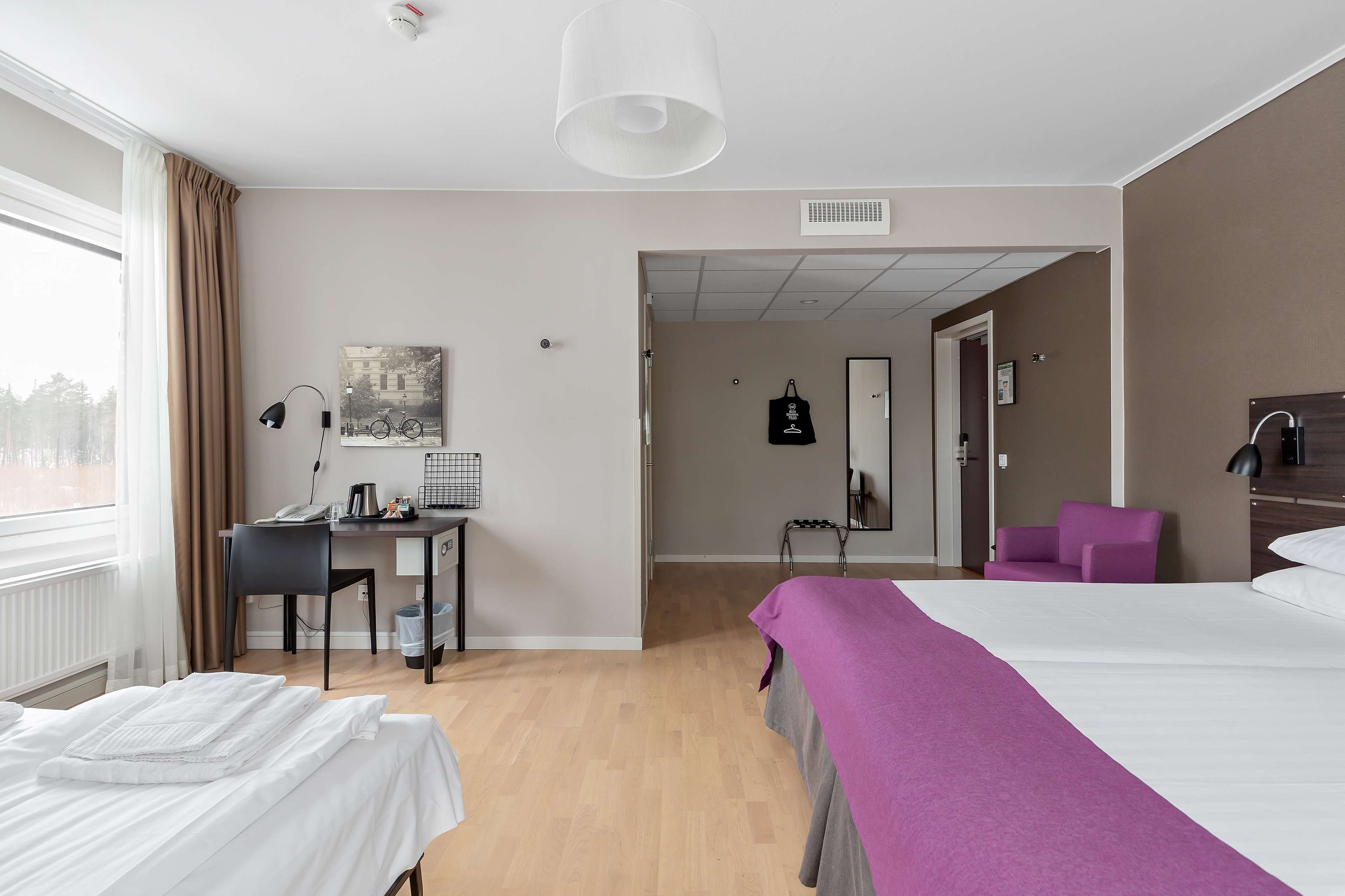 A hotel room with a bed, a desk with a phone, a chair, a purple armchair, and a window with curtains.