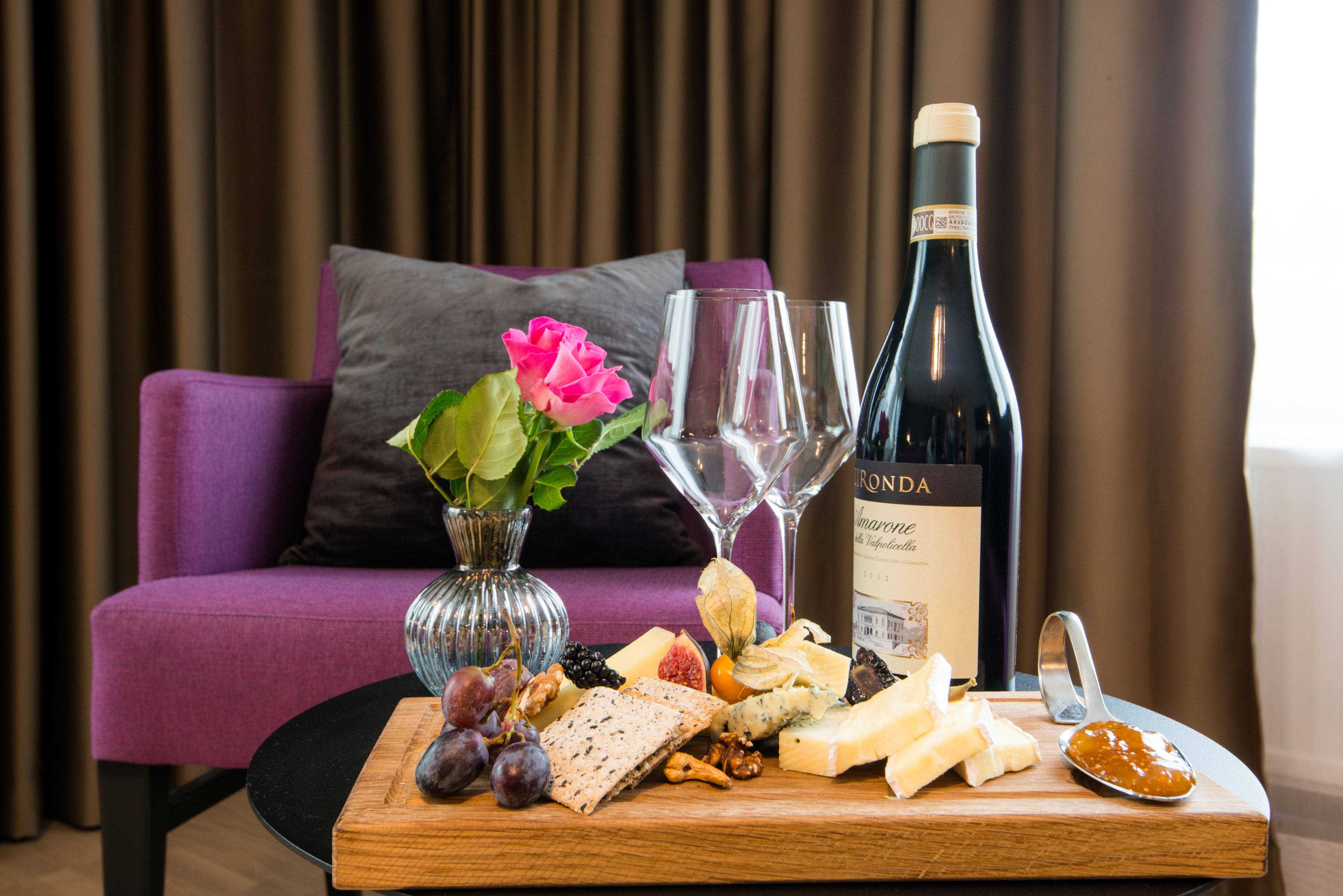 Une table garnie d’une variété de fromages, de fruits, de noix et d’une bouteille de vin, le tout disposé sur une planche en bois devant un canapé violet sur lequel se trouve un vase de fleurs.