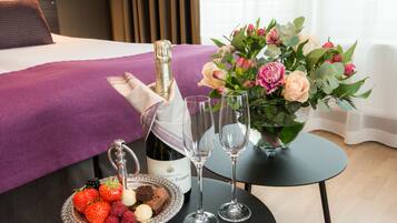 Ein ordentlich eingerichtetes Hotelzimmer mit einem Bett, einem kleinen Tisch, auf dem eine Champagnerflasche, zwei Gläser, eine Obstschale und eine Blumenvase stehen.