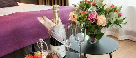 A neatly arranged hotel room with a bed, a small table with a bottle of champagne, two glasses, a bowl of fruit, and a vase of flowers.
