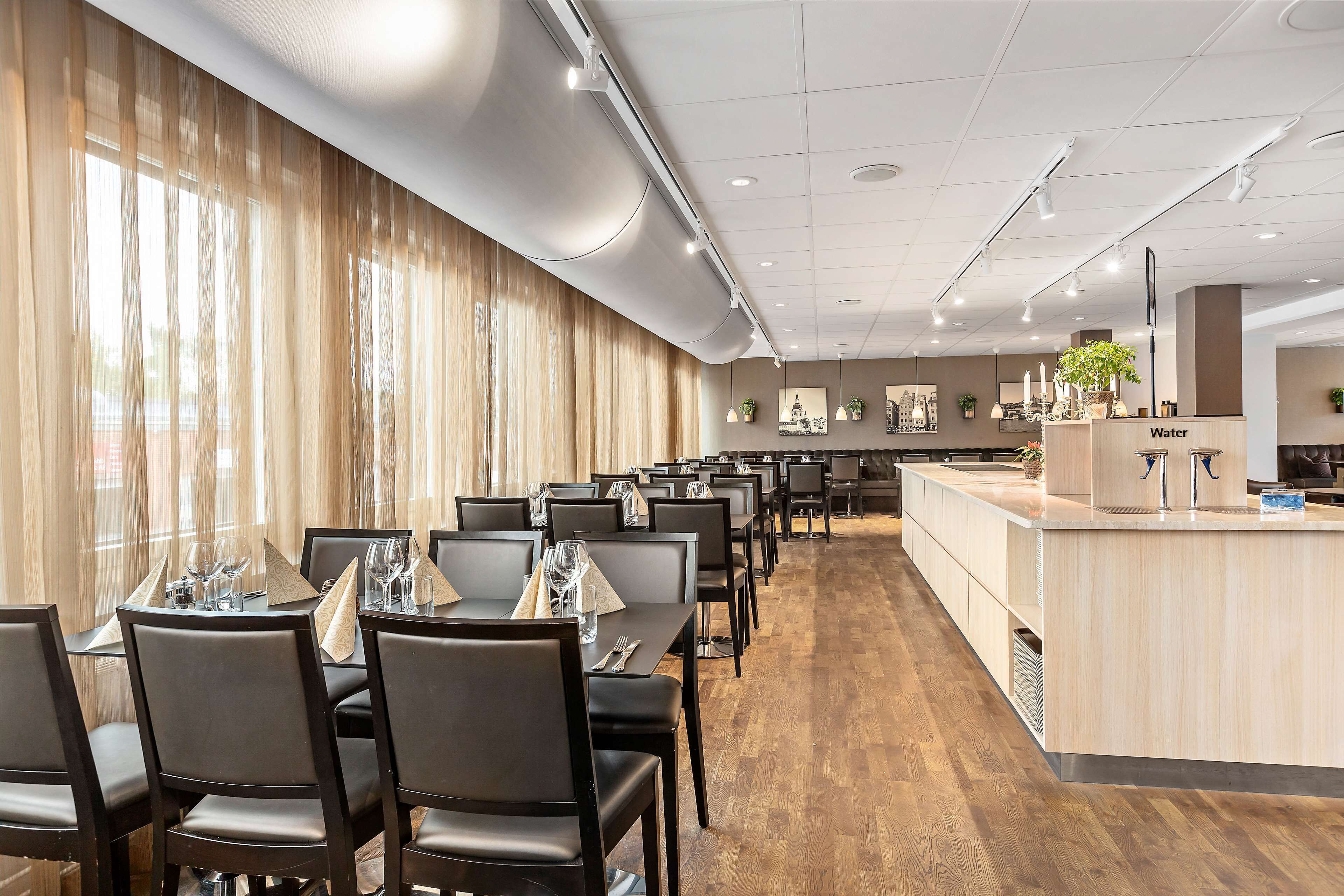 Une salle à manger d’hôtel avec des tables dressées pour le service, un point d’eau et un long comptoir agrémenté d’un miroir et de plantes.