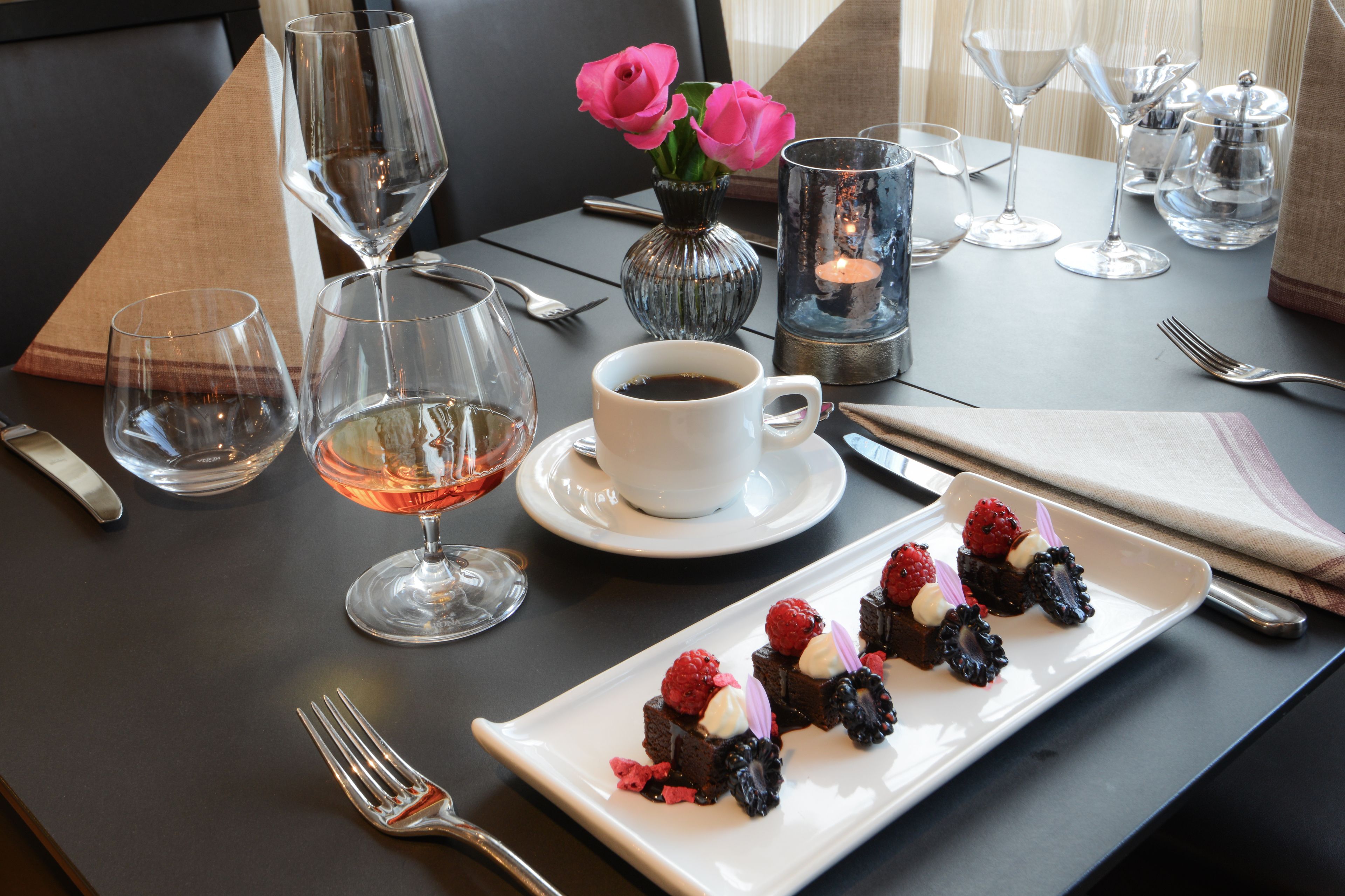 A dining table set with wine glasses, a coffee cup, a vase with pink flowers, and a plate of dessert.