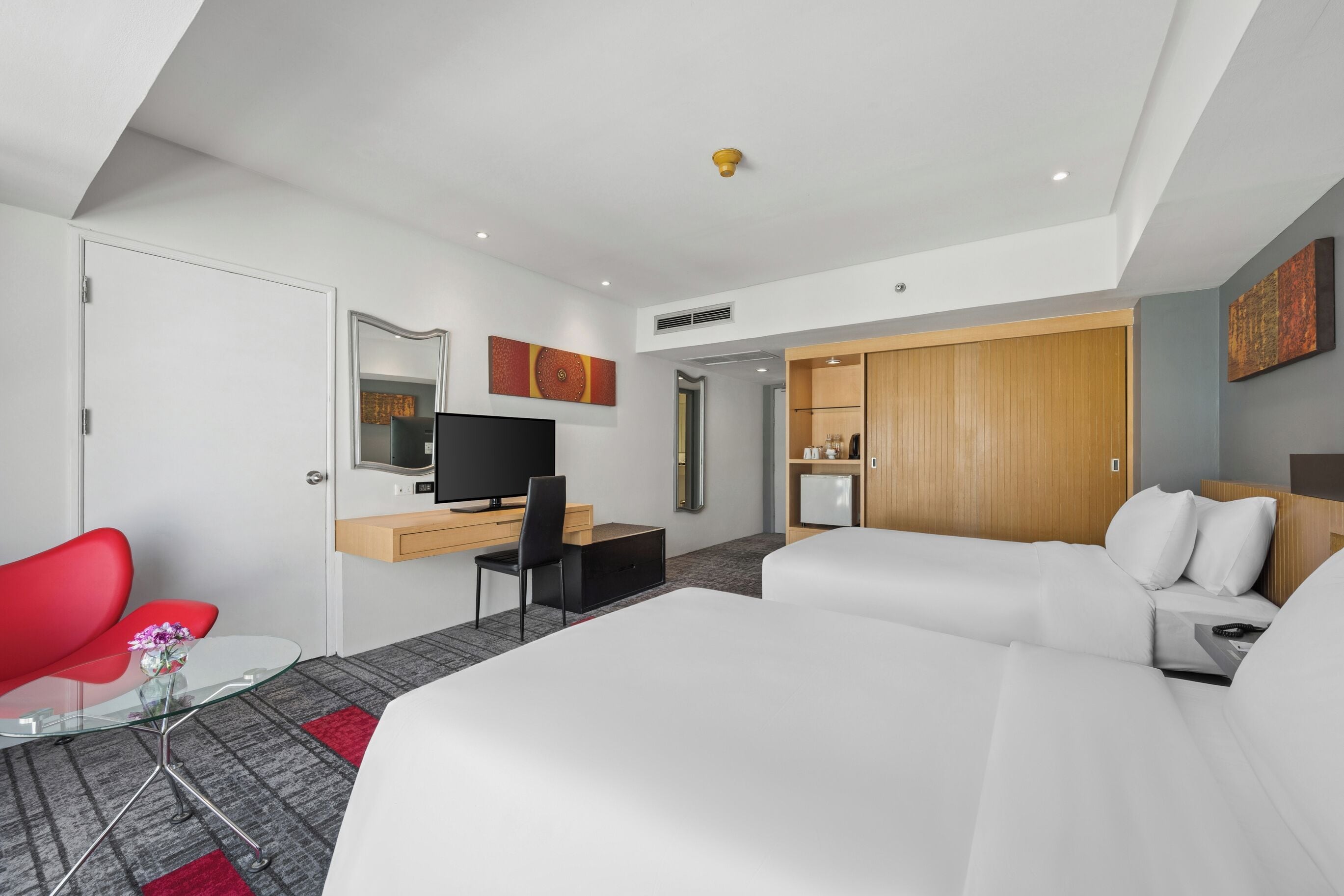 Habitación de hotel moderna con cama, escritorio con televisor, una silla roja y una mesita con flores.