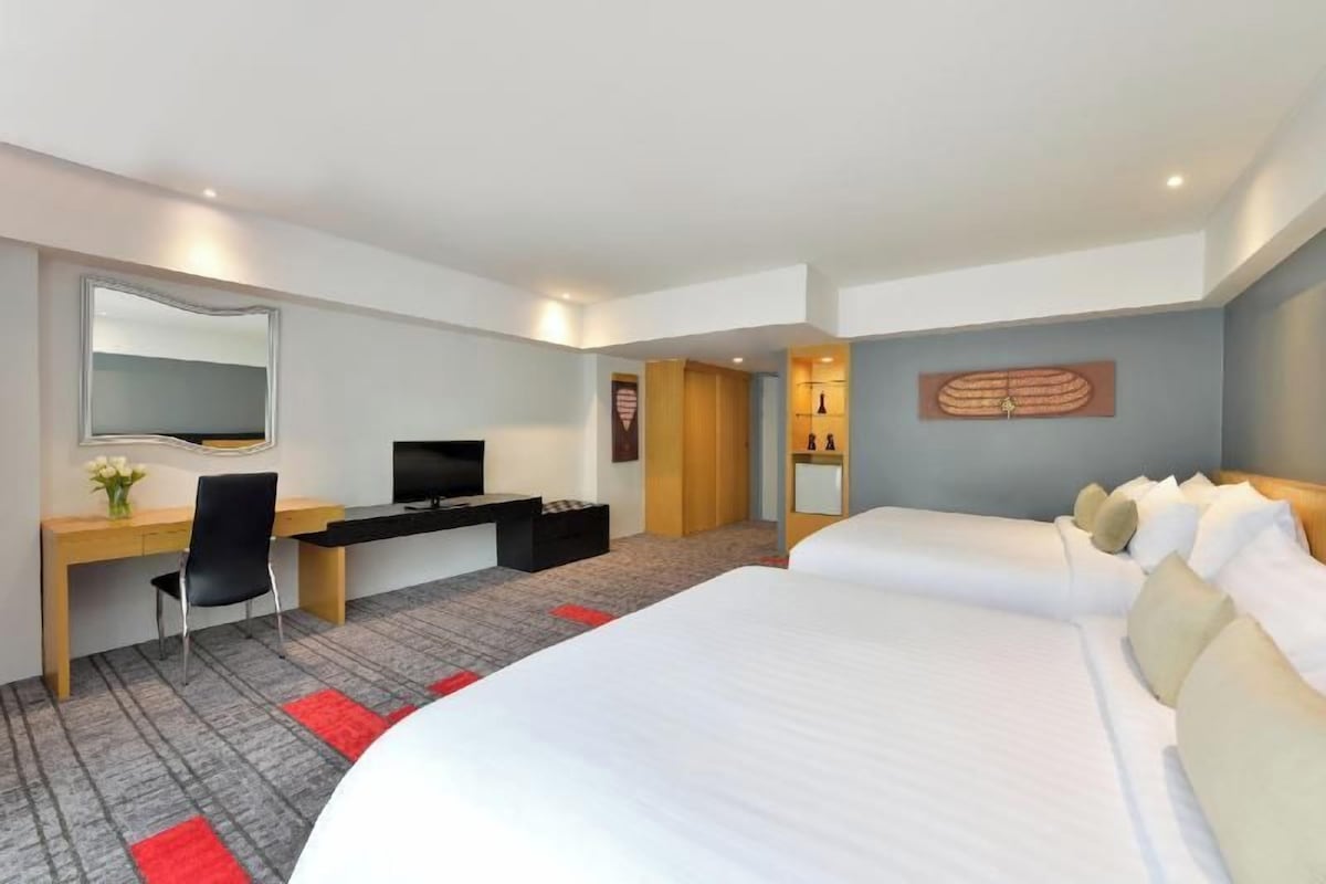 Une chambre d’hôtel comprenant un lit, un bureau avec une chaise, une télévision, un miroir et une petite table sur laquelle est posé un vase de fleurs.