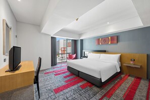 Une chambre d’hôtel avec un lit, un bureau avec une télévision, une chaise rouge, un balcon donnant sur des immeubles et un tapis rouge et gris.