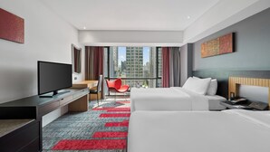A hotel room with a TV, desk, red chair, two beds, and a view of tall buildings. - BelAire Bangkok Sukhumvit (Bangkok)