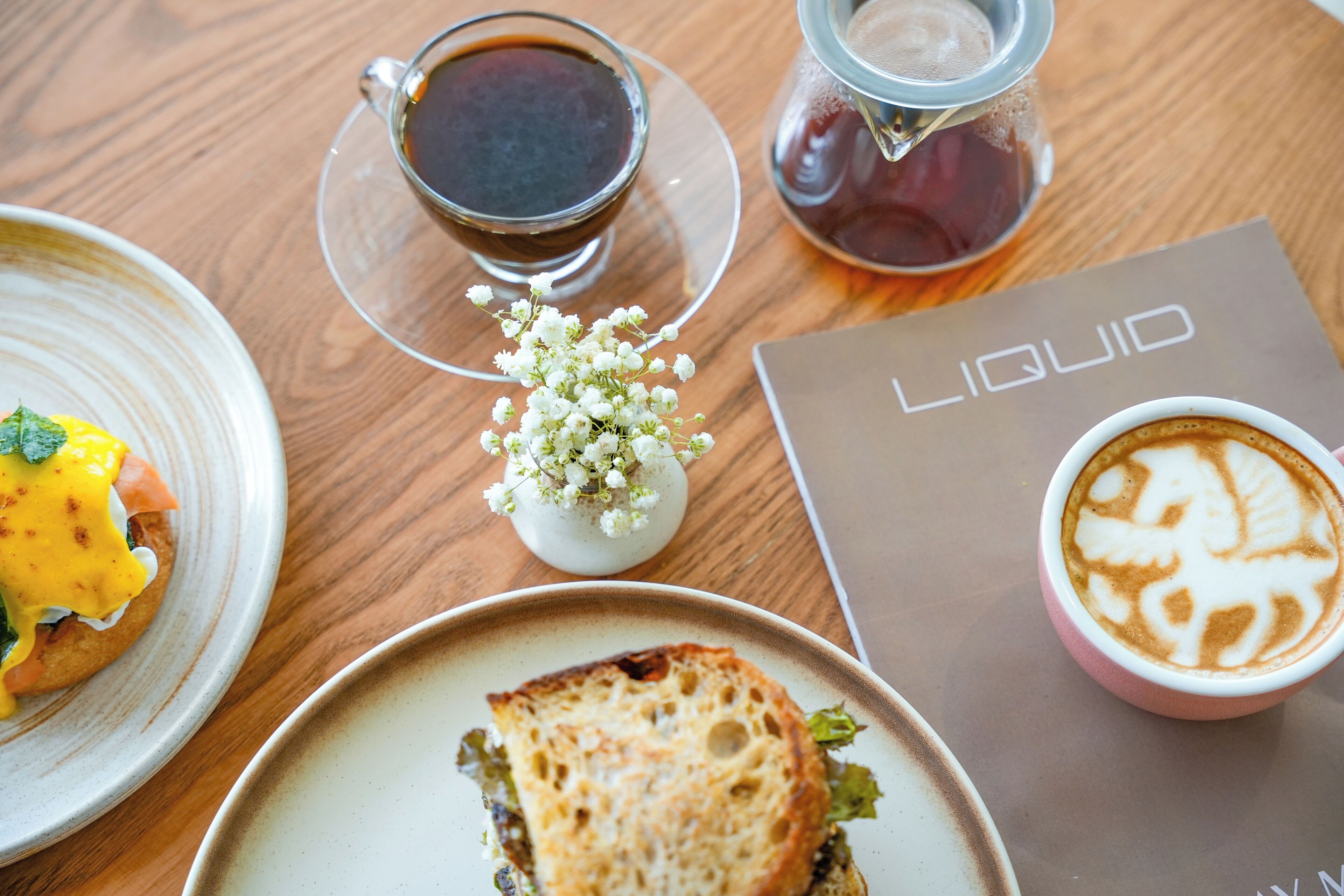 Una mesa con un sándwich, una taza de café, una jarra de té y un folleto titulado "LIQUID".