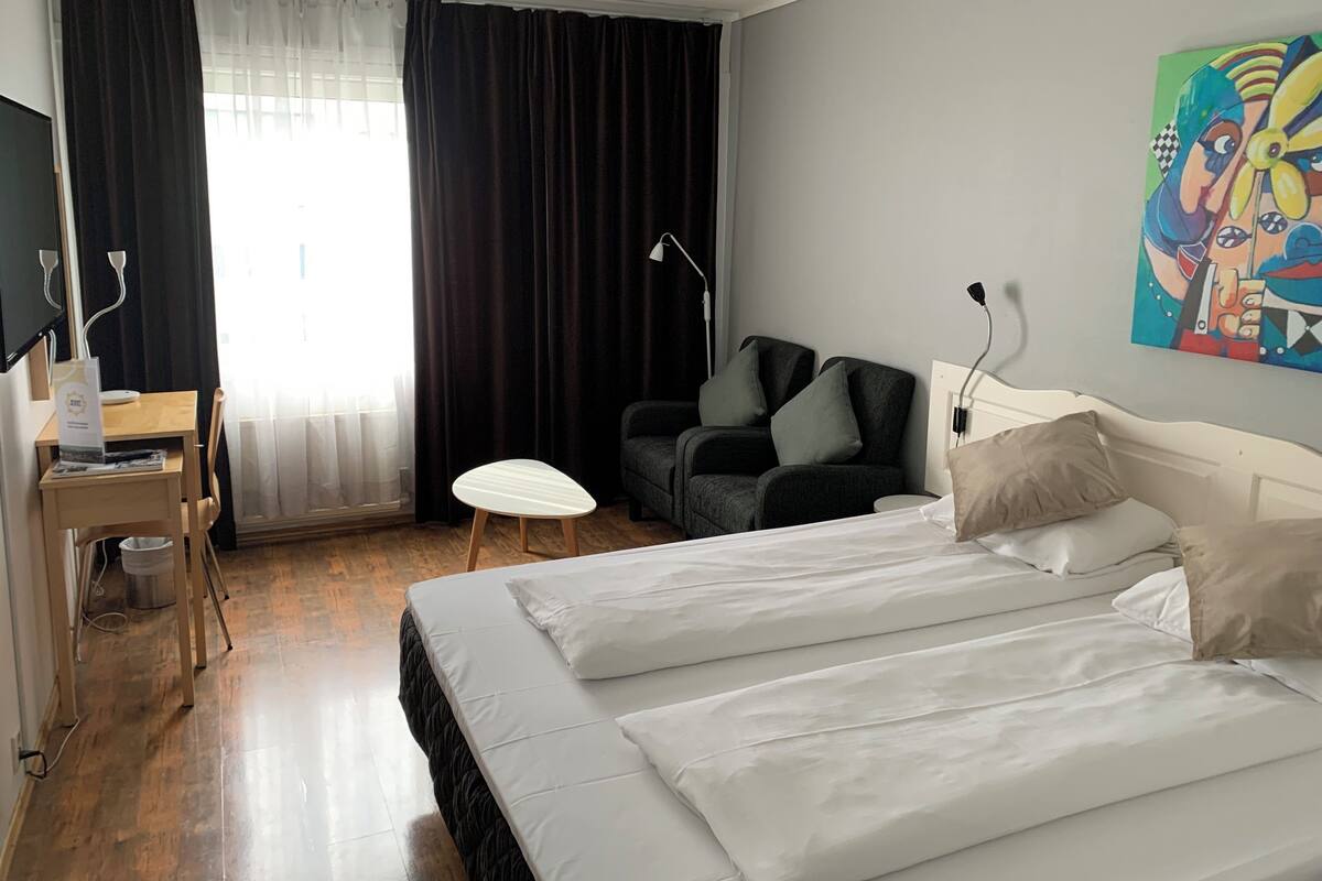A hotel room with a bed, a desk, a chair, a small table, and a painting on the wall.