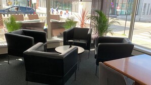 Un espace de détente intérieur moderne, avec des canapés en cuir et une table en bois, de grandes fenêtres donnant sur la rue et des plantes en pot.