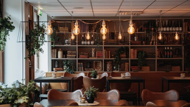 Ein gemütliches Restaurant mit Holztischen und -stühlen, einem Bücherregal voller Bücher und Flaschen sowie hängenden Glühbirnen.