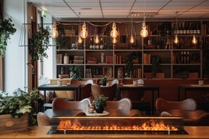 A cozy restaurant with wooden tables and chairs, a bookshelf filled with books and bottles, and hanging light bulbs. - Home Hotel Slottsparken (Linkoping)