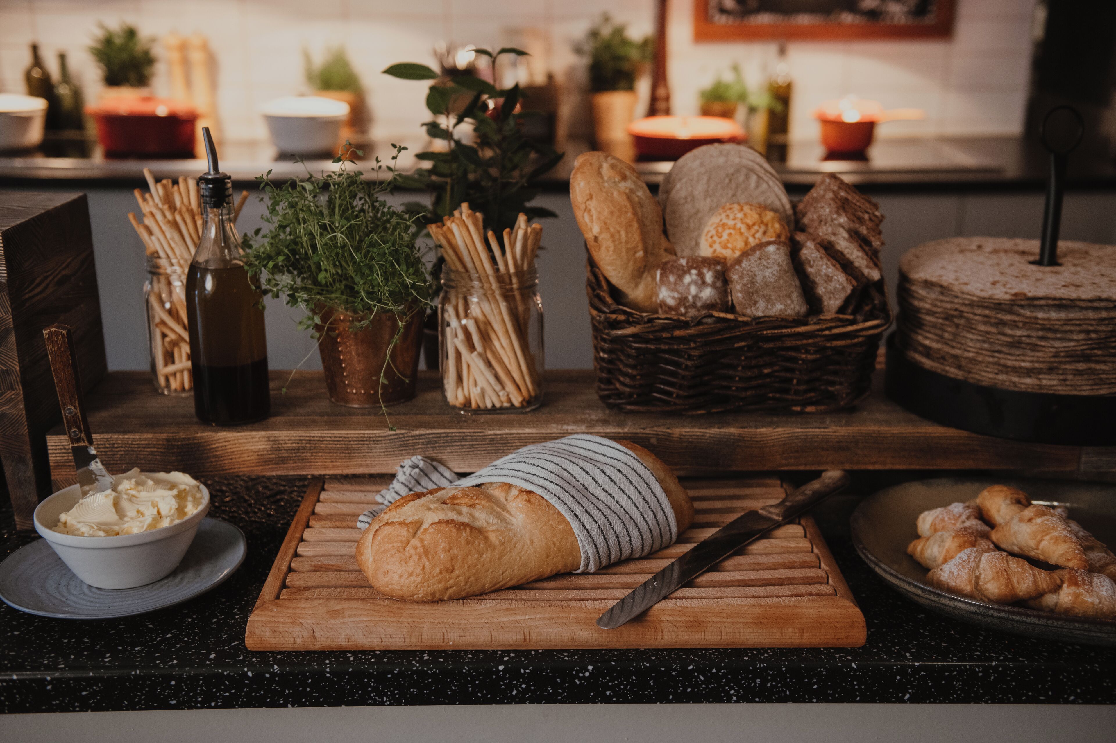 A kitchen counter with various breads, a cutting board, a knife, and a bowl of butter.