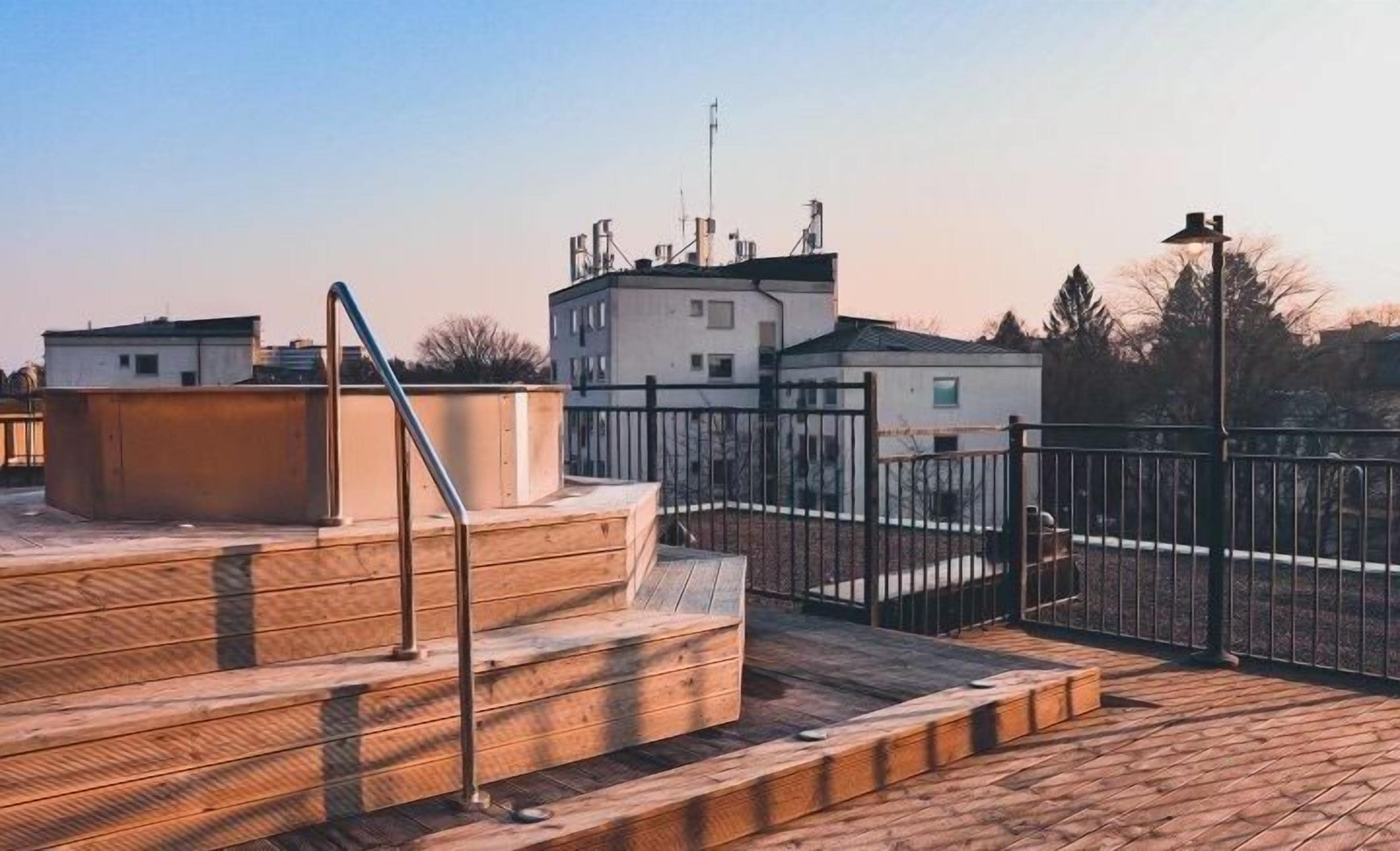 Terraza en la azotea con escalones de madera y barandillas de metal, rodeada por una cerca de metal.