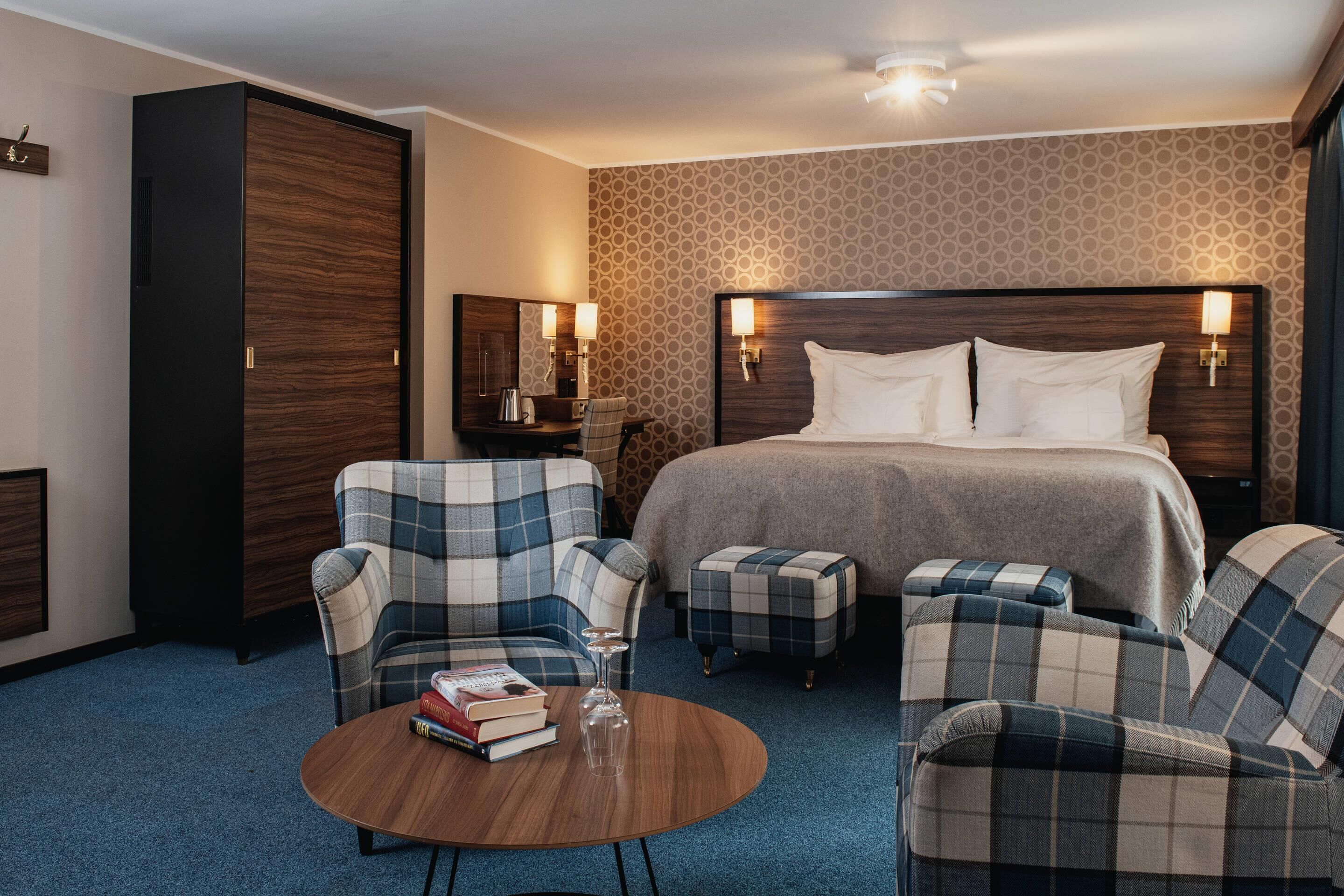 A hotel room with a large bed, two armchairs, a small table with books and a glass, and a wardrobe.