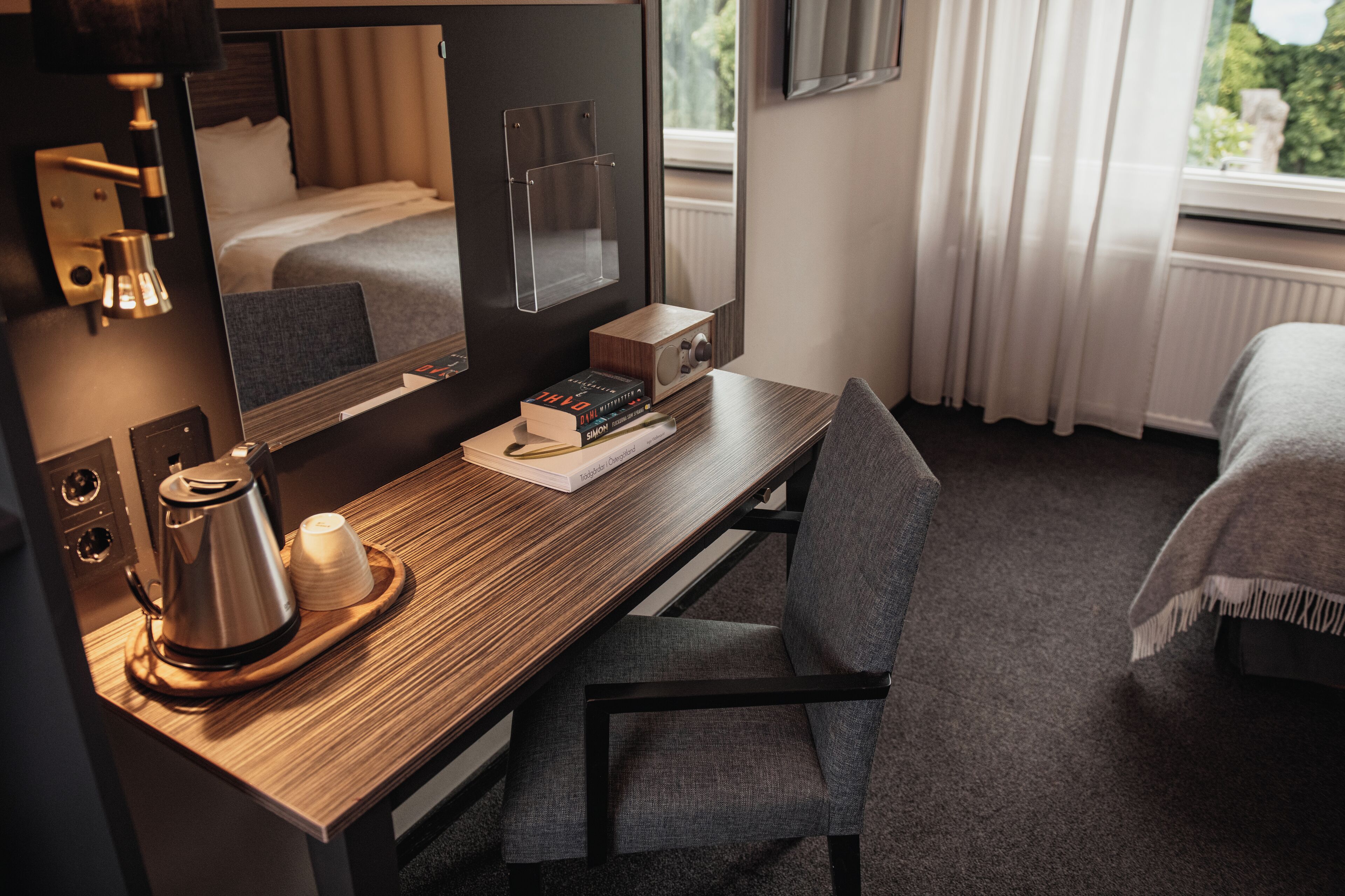 A hotel room with a wooden desk, a chair, a bed, a mirror, a coffee maker, and a window with curtains.