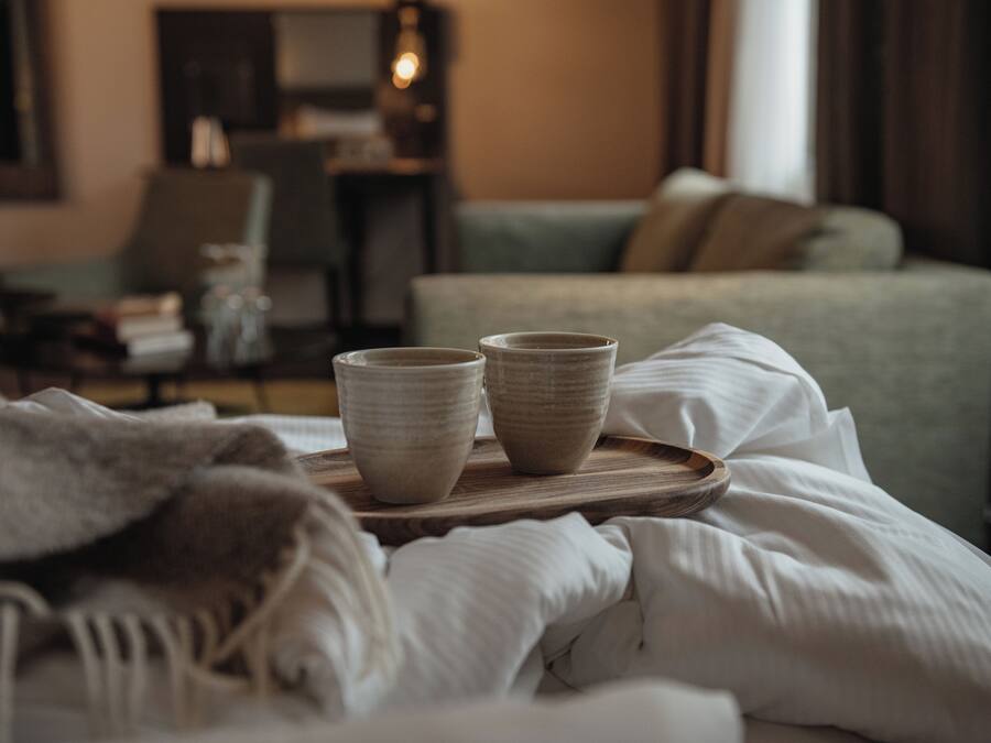Una cama bien tendida con una bandeja que sostiene dos tazas de cerámica, un sofá verde y una silla en una habitación de hotel.