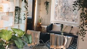 Ein gemütliches Wohnzimmer mit einer Couch, Kissen und einem Couchtisch. Mehrere Blumentöpfe und eine hängende Pflanze sind ebenfalls vorhanden.