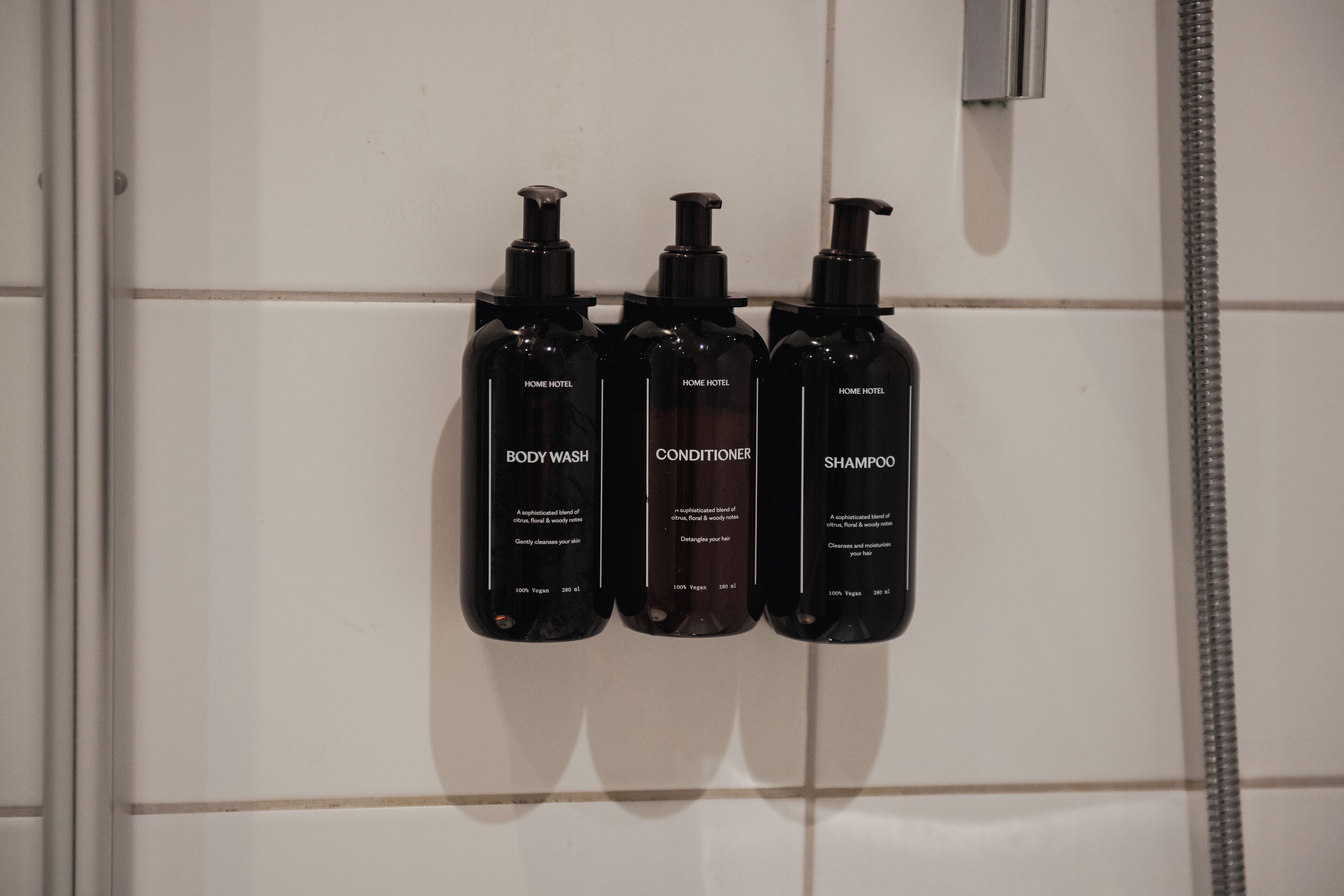 Drie zwarte flesjes toiletartikelen op een betegelde muur.