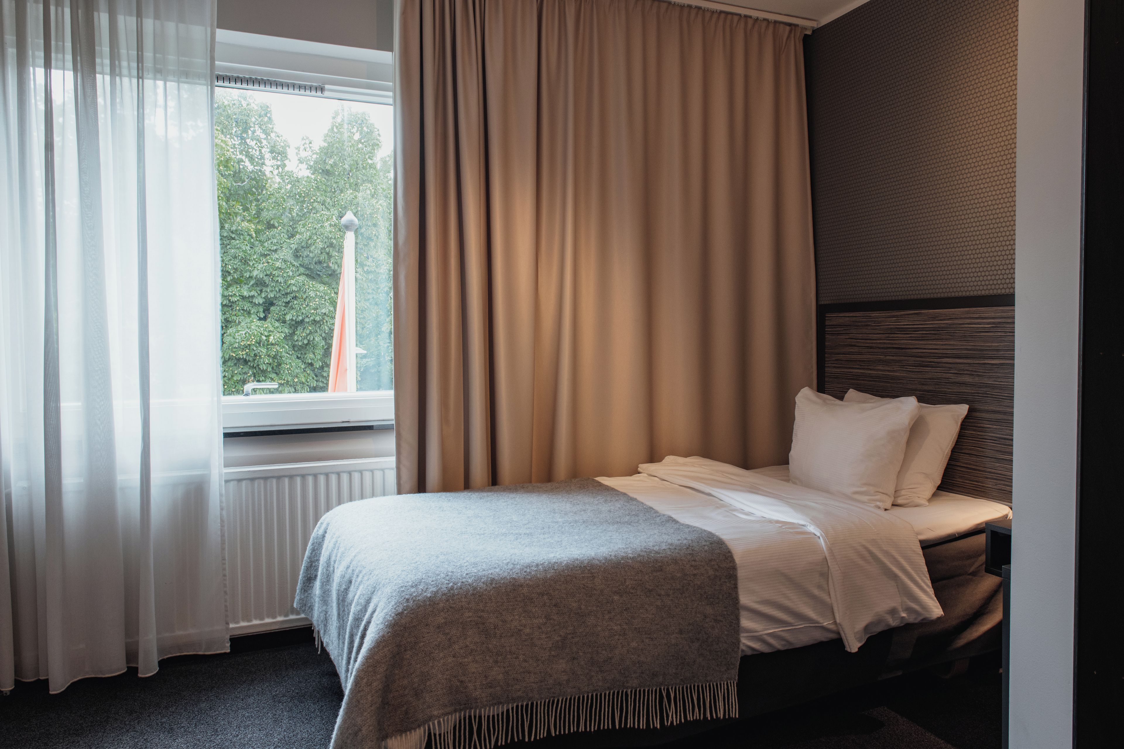 Habitació d'hotel amb un llit, una finestra amb cortines i vistes a la vegetació.