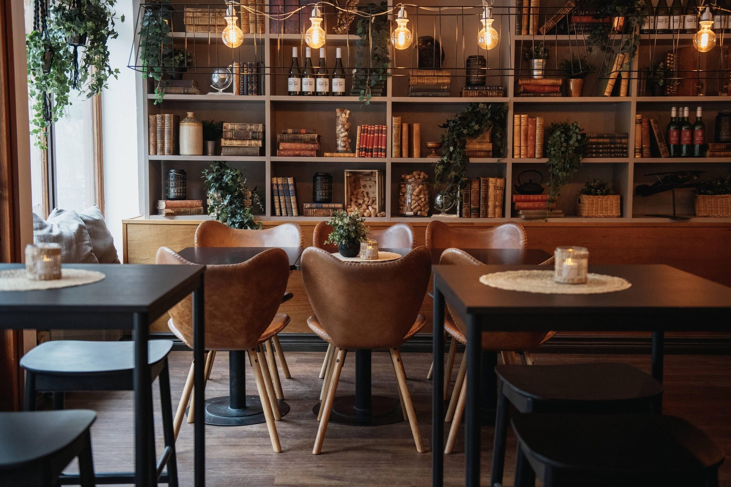 Un accogliente caffè con tavoli e sedie in legno, una libreria piena di libri e oggetti decorativi, e lampadine a sospensione.