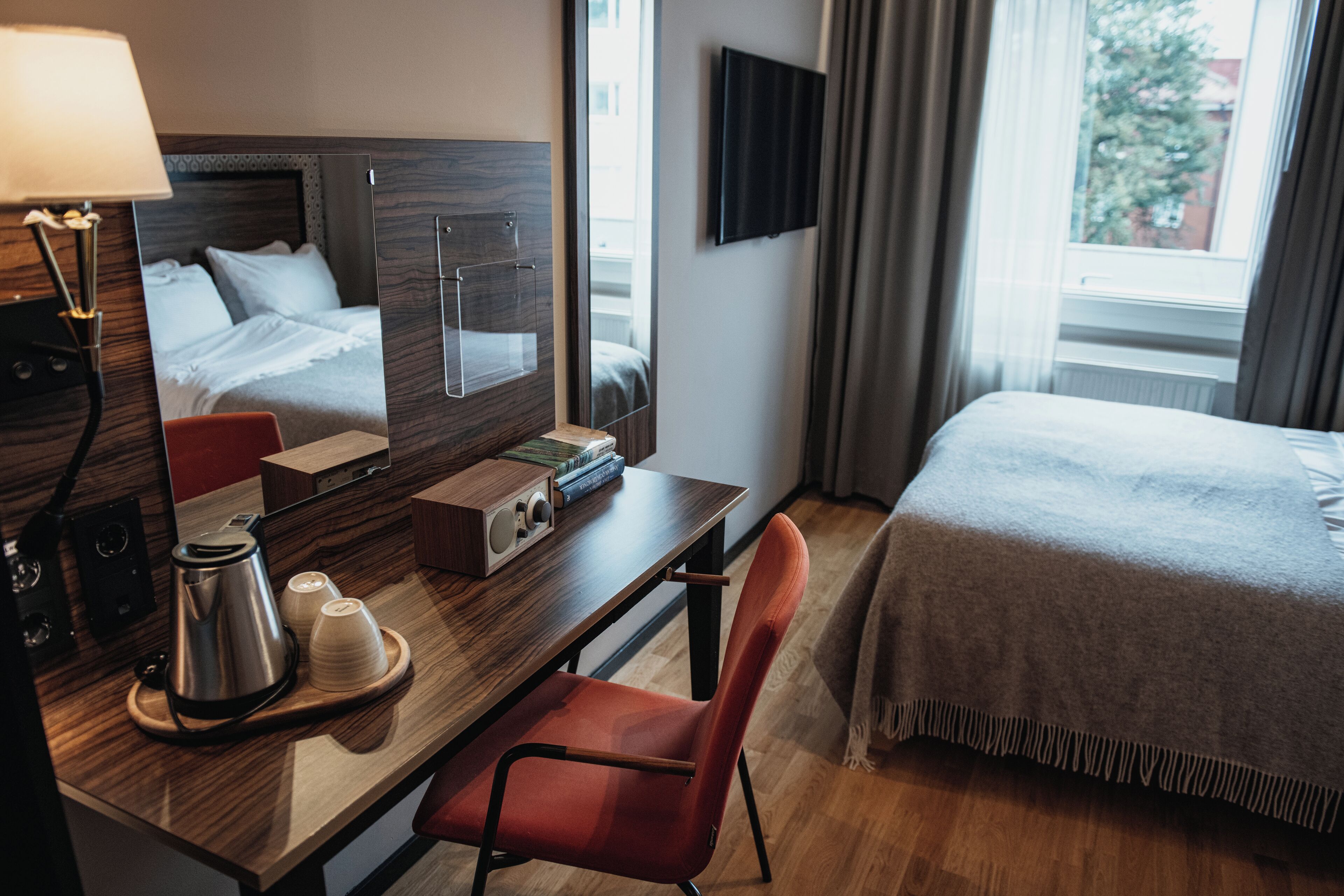 A hotel room with a bed, a desk with a coffee maker, a chair, a TV, and a window with curtains.