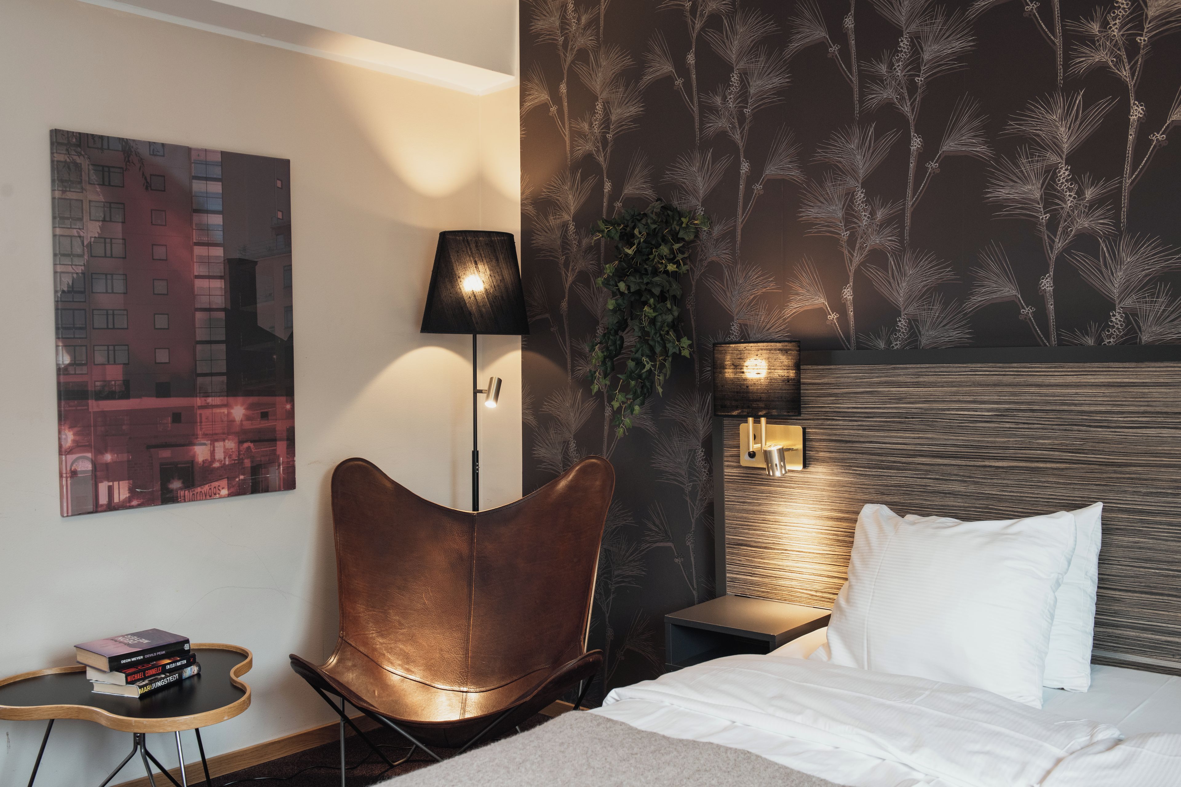 A hotel room with a bed, a brown leather chair, a small table with books, a wall-mounted lamp, and a cityscape painting.