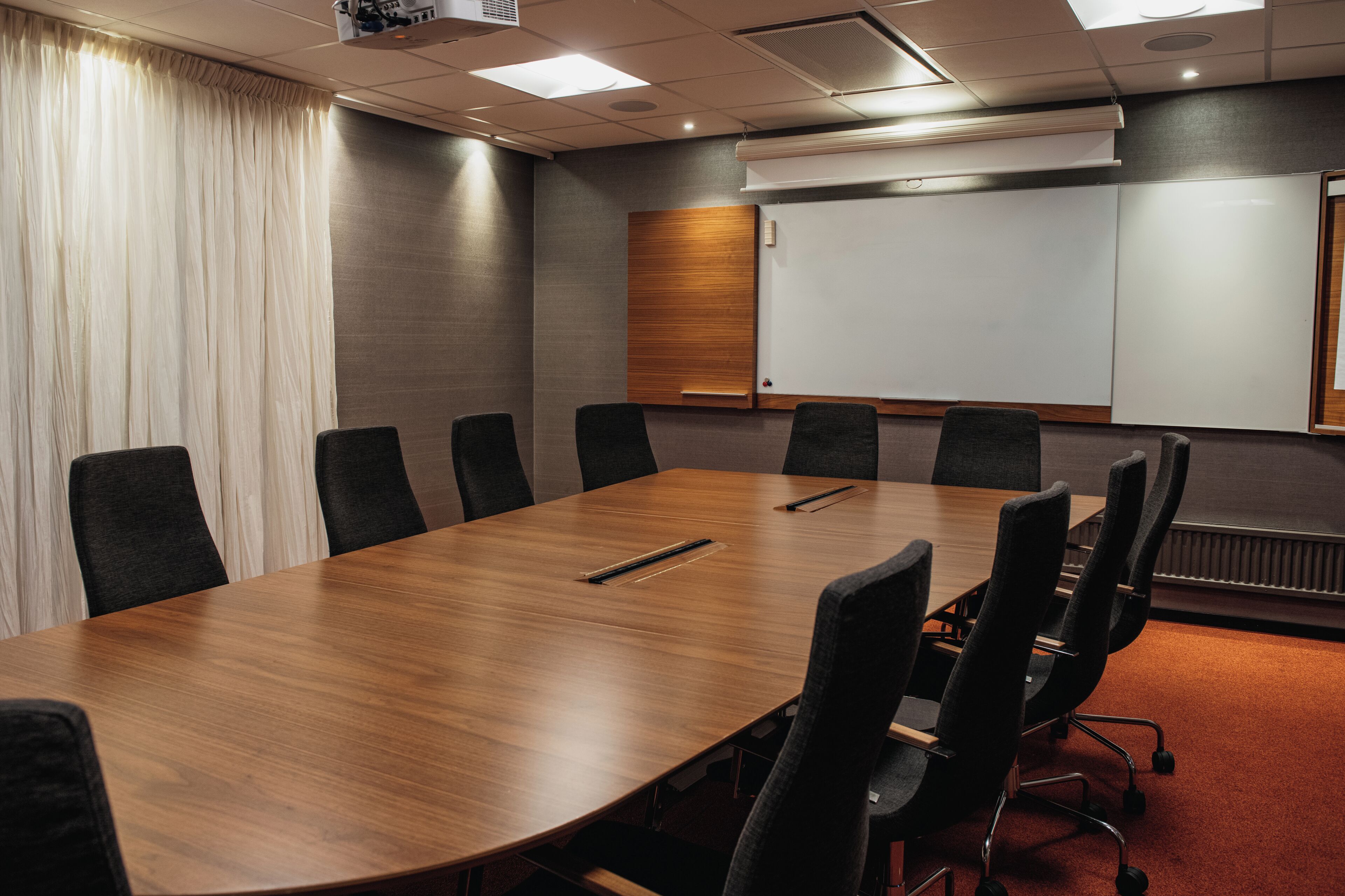 A conference room with a long wooden table, black chairs, a whiteboard, and a projector.