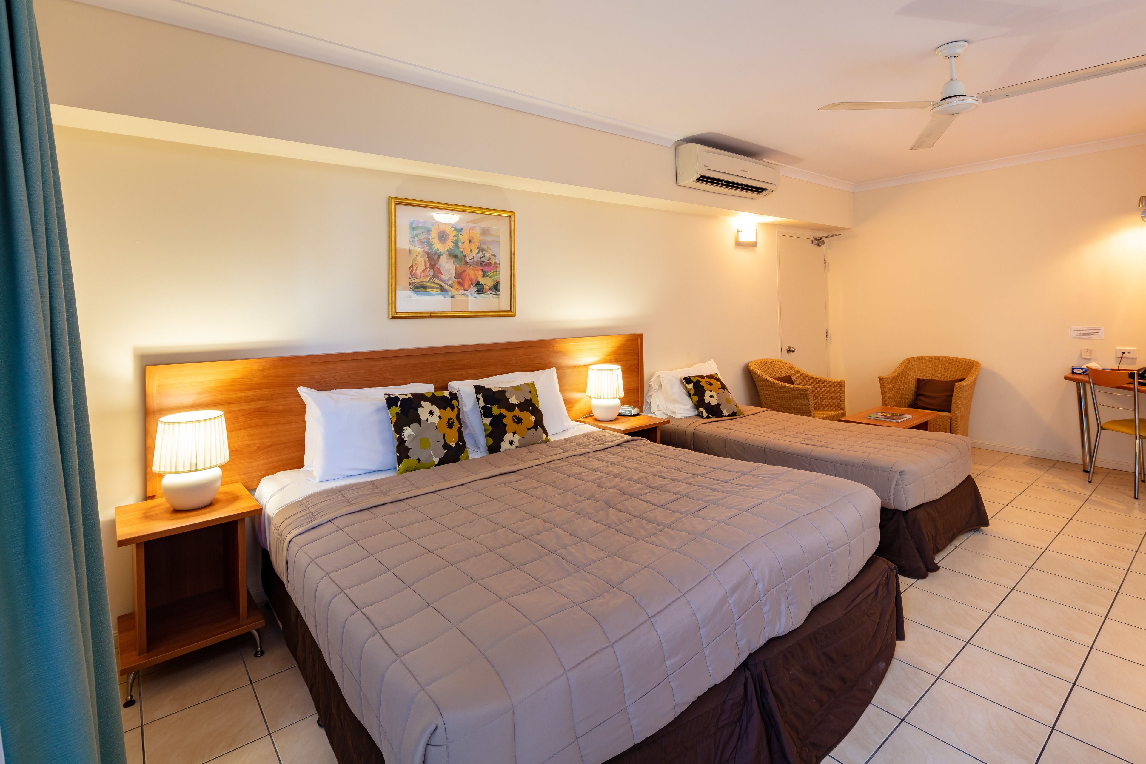 A hotel room with two beds, a wooden headboard, a ceiling fan, an air conditioning unit, a small table, and a chair.