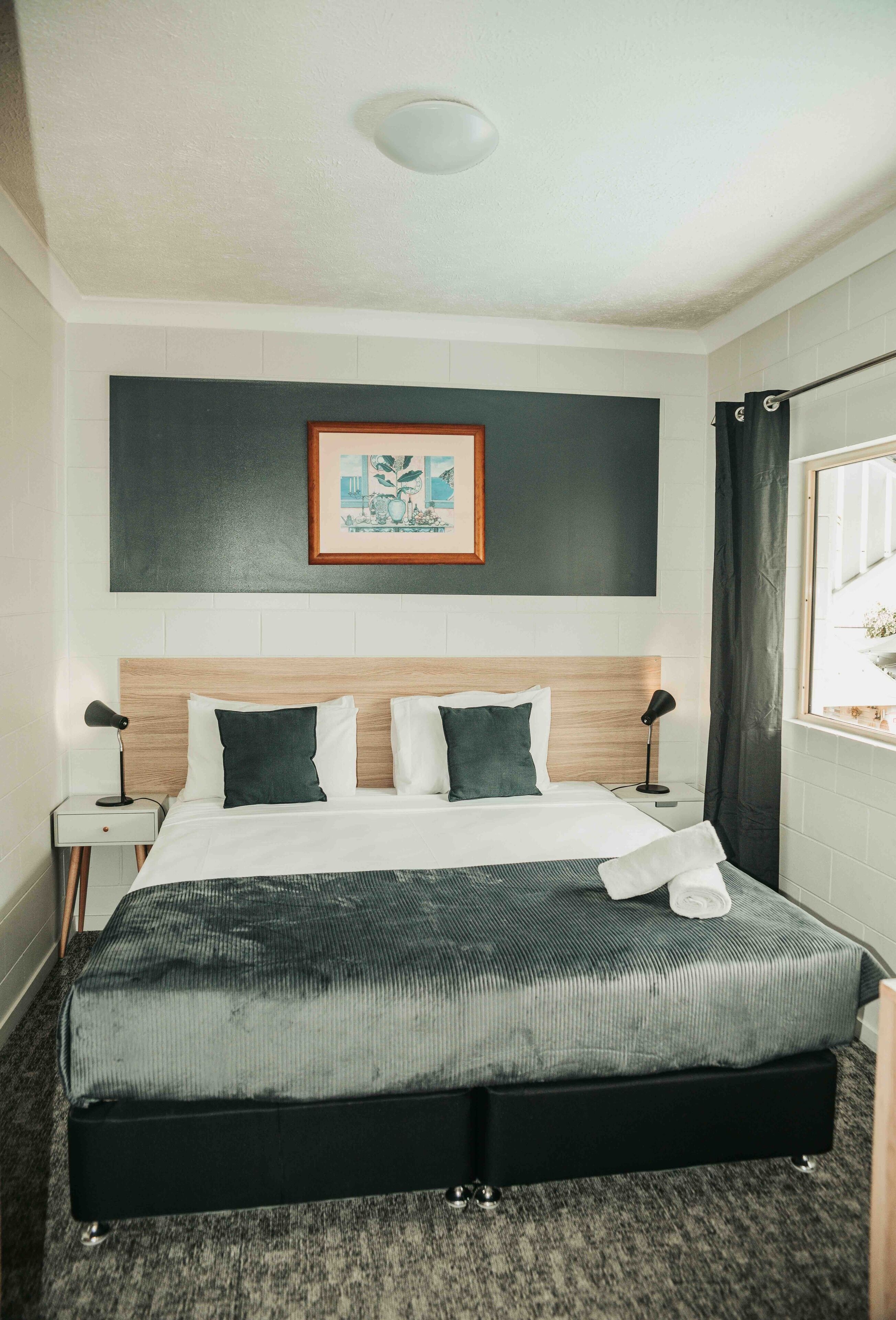 A hotel room with a large bed, a framed picture above the bed, a window with a view, and a nightstand with a lamp.