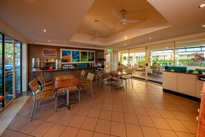 A spacious indoor dining area with multiple tables and chairs, a counter with a cash register, and a view of the outdoors through large windows. - Cairns Queens Court (Cairns North)
