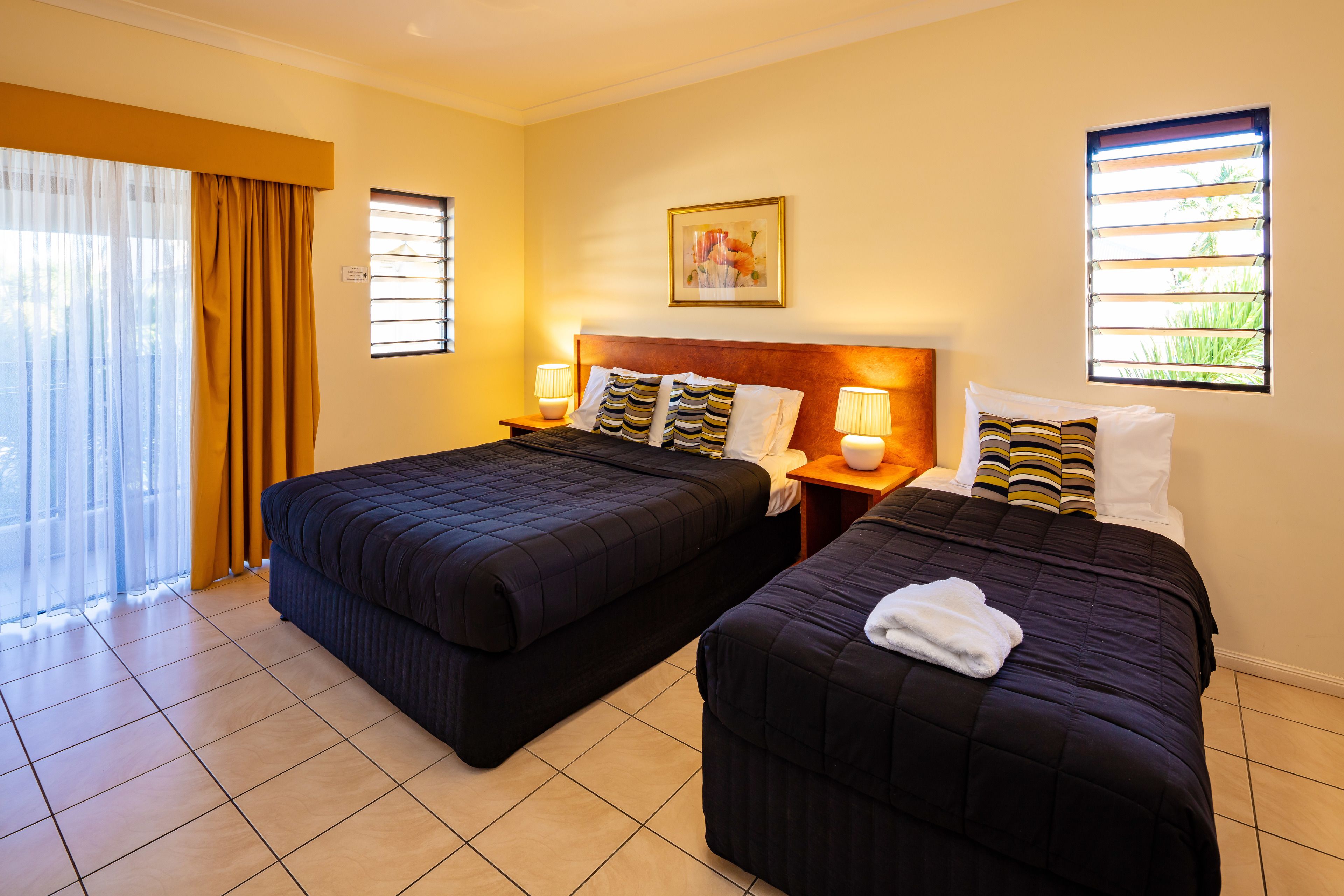 A hotel room with two beds, a large window with blinds, a painting on the wall, and a view of greenery outside.