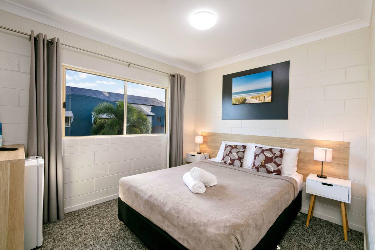 Une chambre d’hôtel avec un grand lit, une fenêtre avec des rideaux, une table de chevet avec une lampe et un tableau au mur.