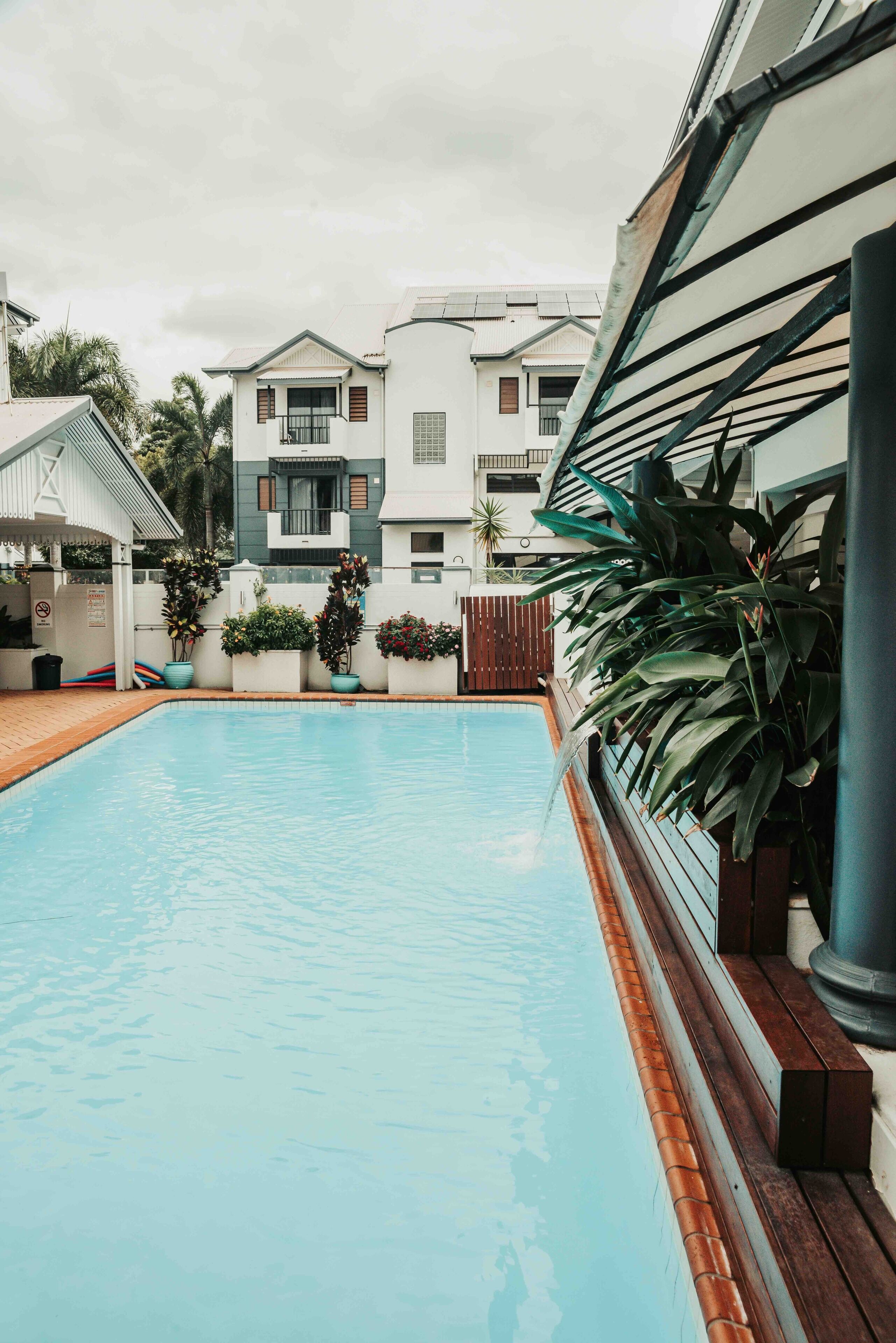 A swimming pool with a wooden deck, surrounded by potted plants and a white building with balconies.