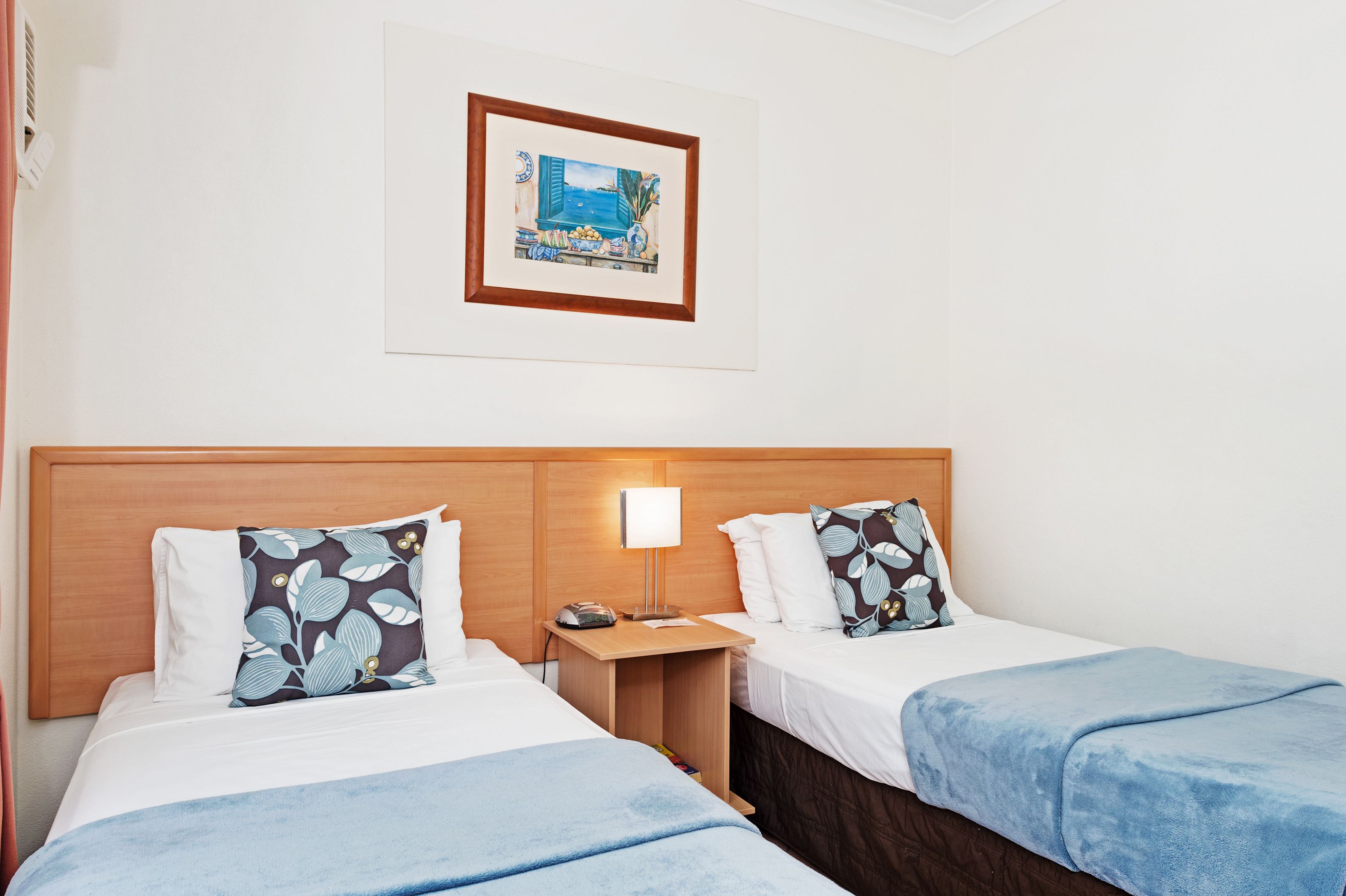 A hotel room with two beds, a wooden headboard, a bedside table with a lamp, and a framed picture on the wall.