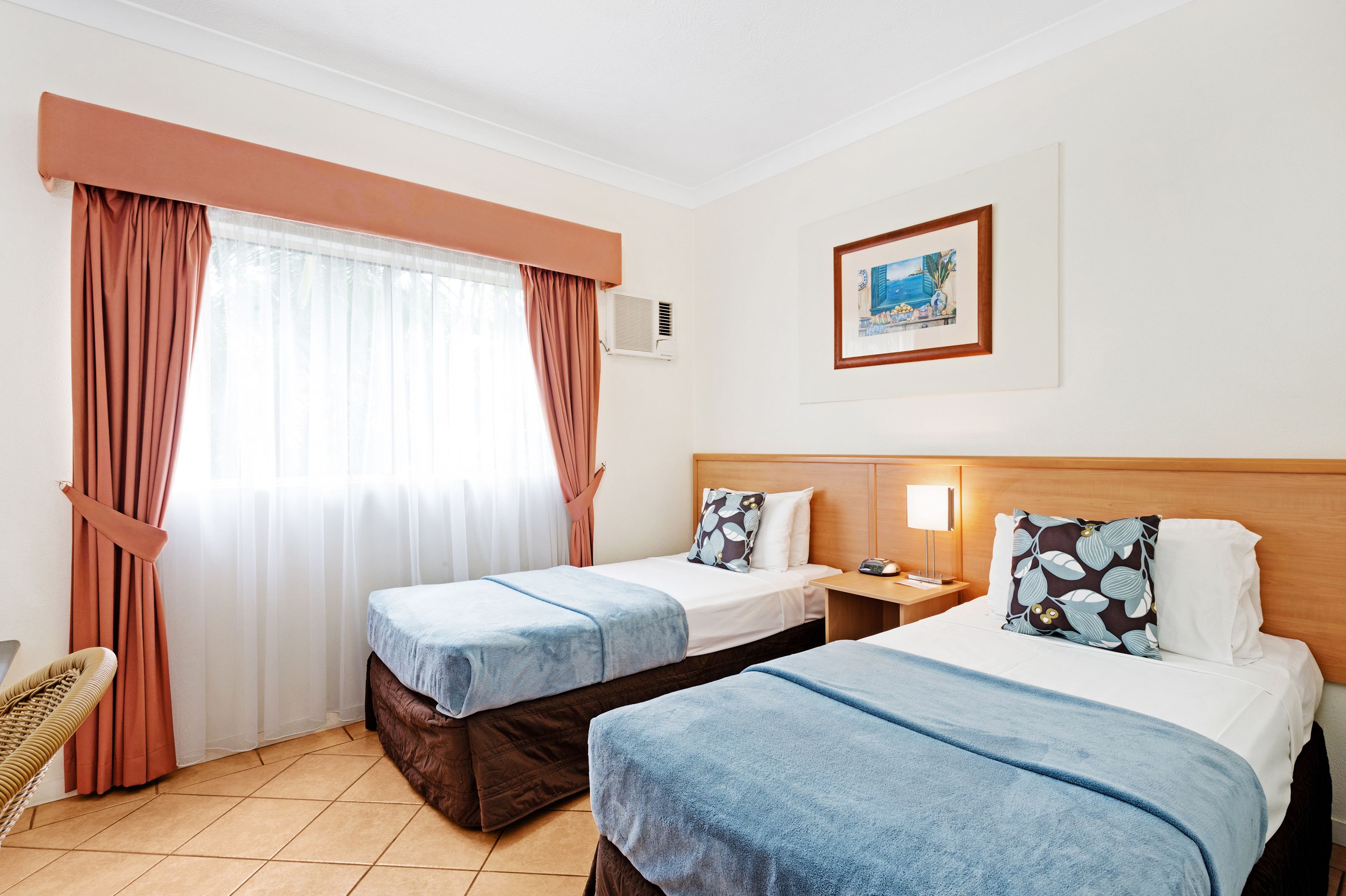 A hotel room with two beds, a chair, a painting on the wall, and a window with curtains.