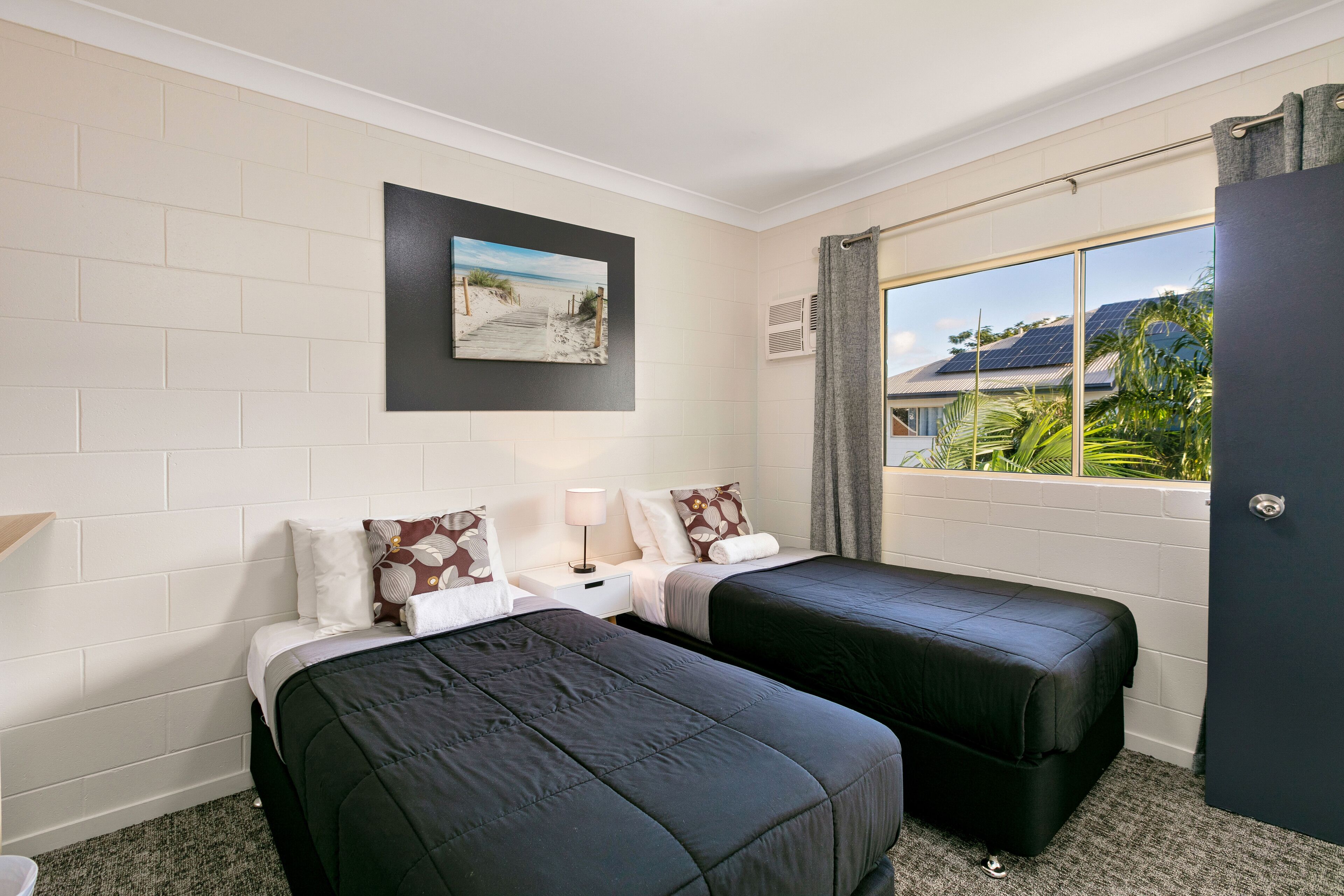 A hotel room with two beds, a nightstand, a lamp, a picture on the wall, and a window with a view of greenery and a building.