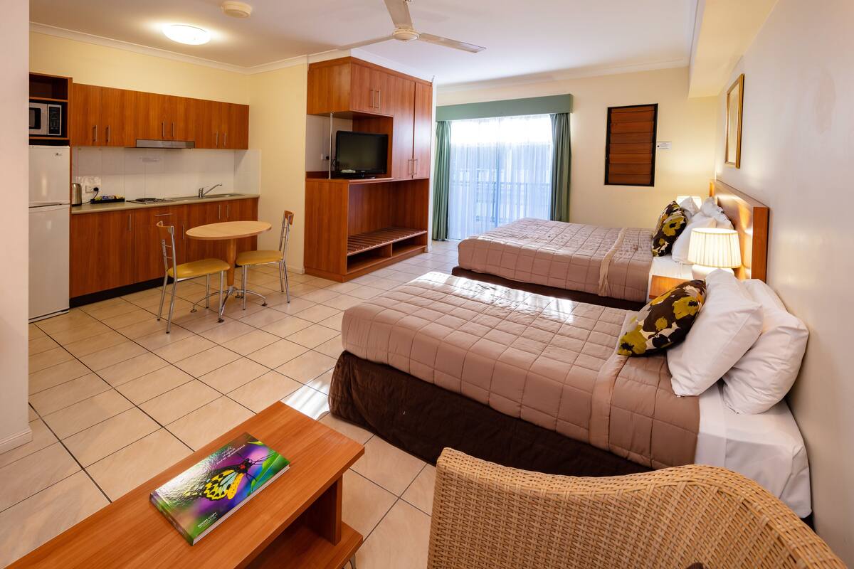 Une chambre d’hôtel compacte comprenant un coin cuisine, deux lits, un espace repas et une grande fenêtre avec des rideaux.