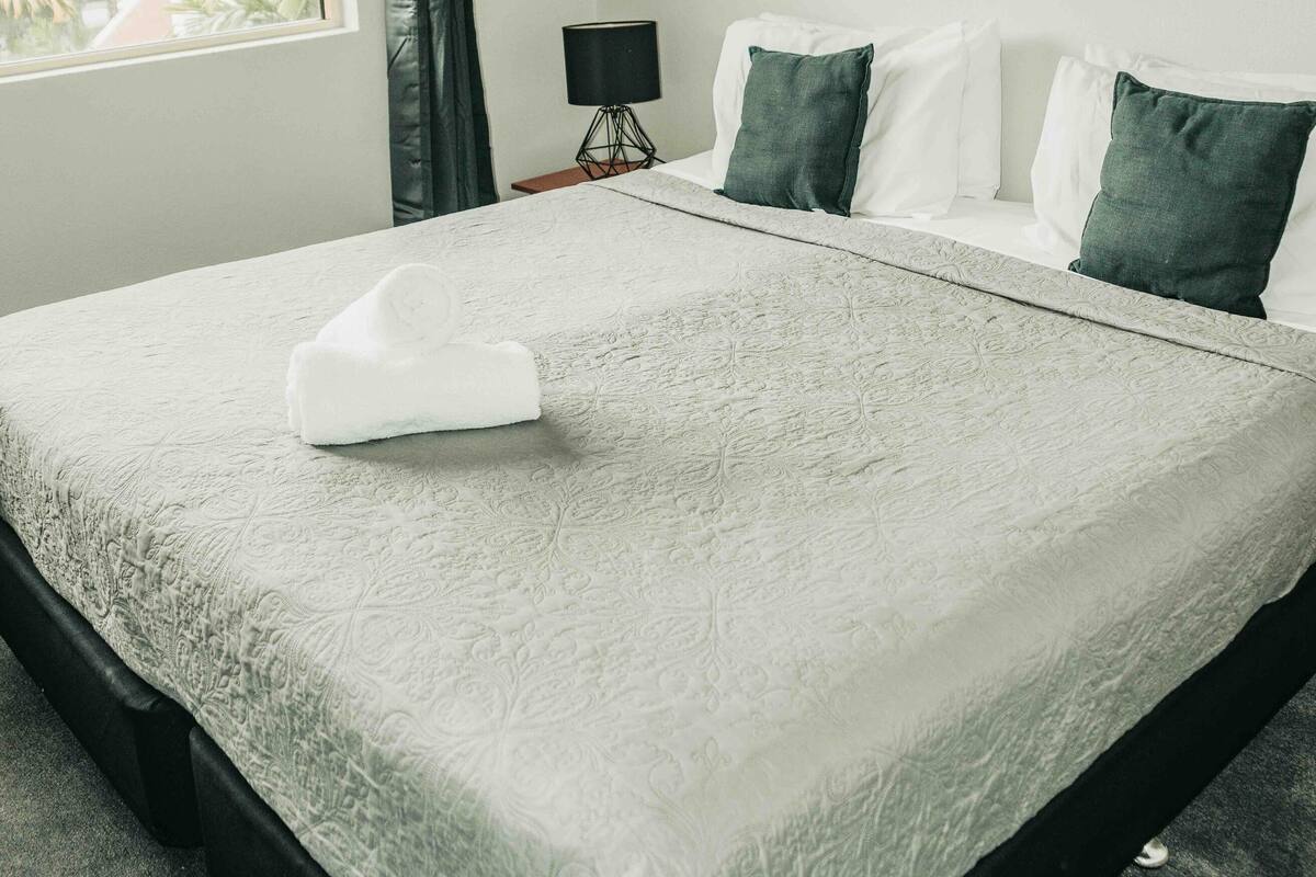 Un lit bien fait, avec une couette blanche et une serviette pliée, une œuvre d’art encadrée au mur et une fenêtre avec des rideaux.