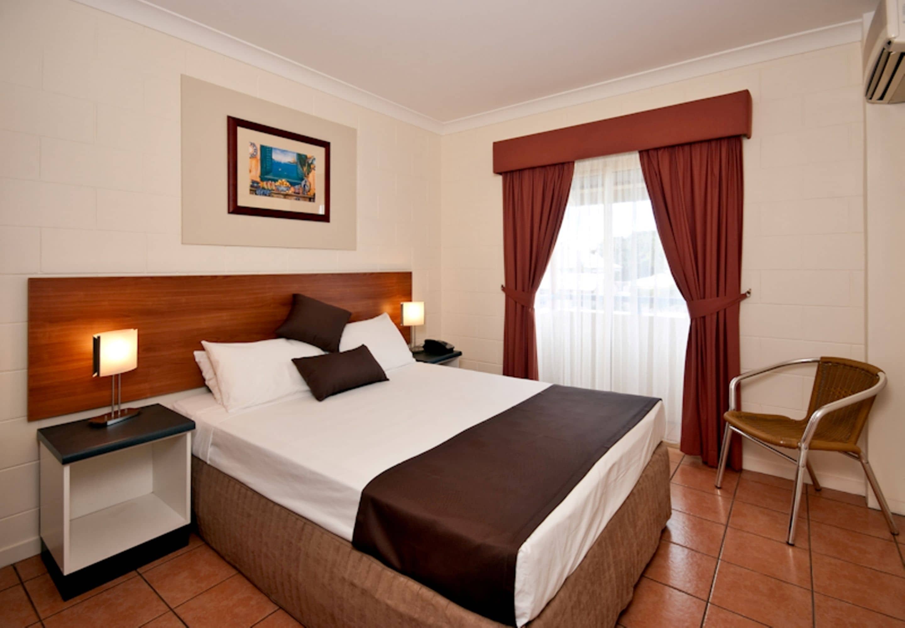Habitación de hotel con cama, mesitas de noche, una silla, ventana con cortinas e imagen en el marco en la pared.