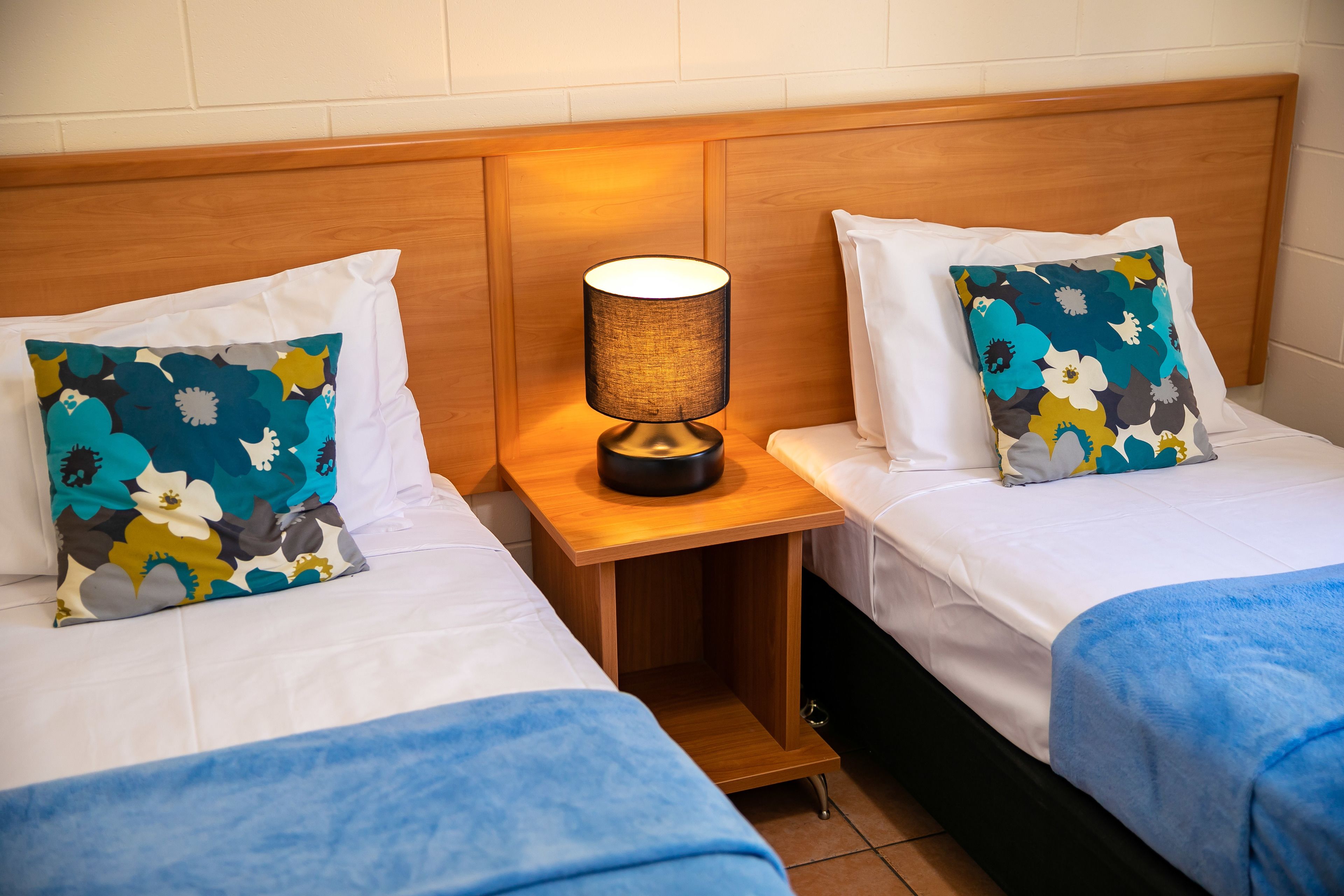 Two single beds with white linens, a wooden nightstand with a lamp, and floral-patterned pillows.