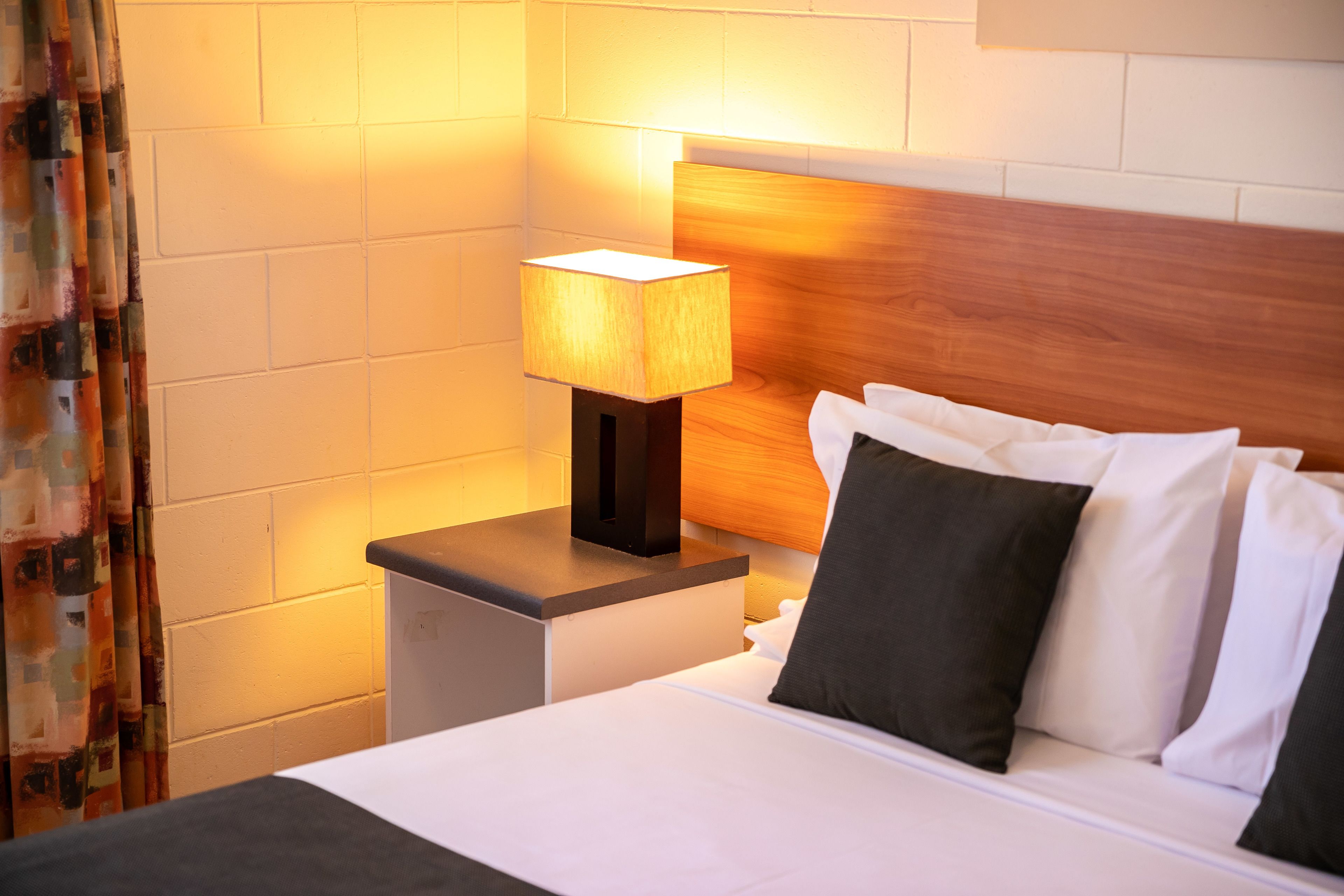 Ein ordentlich bezogenes Bett mit weißen und schwarzen Kissen, einem Holz-Kopfteil und einer Nachttischlampe auf einem kleinen Beistelltisch.