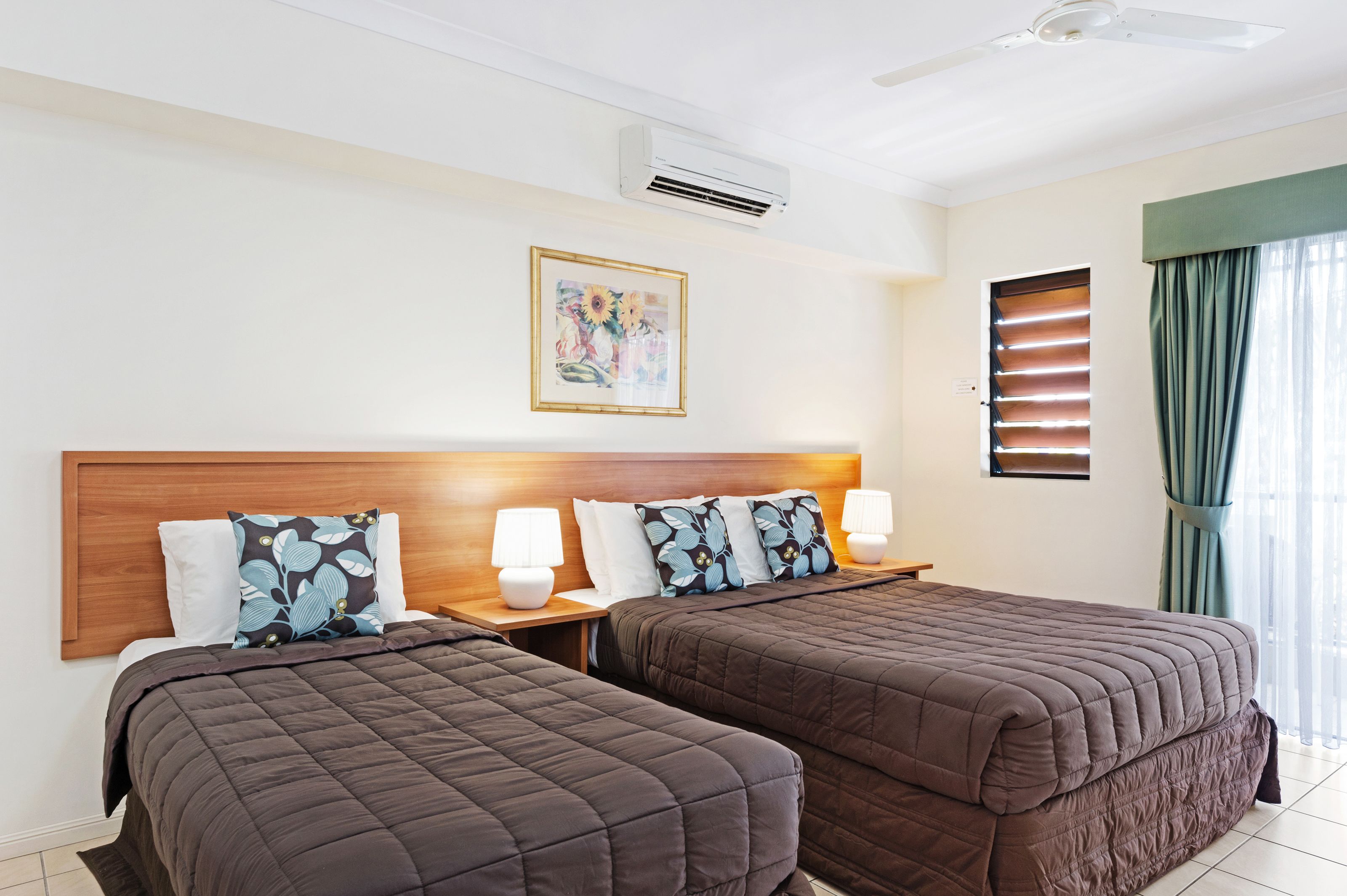A hotel room with two beds, a wooden headboard, a ceiling fan, an air conditioner, a window with blinds, and a framed picture on the wall.
