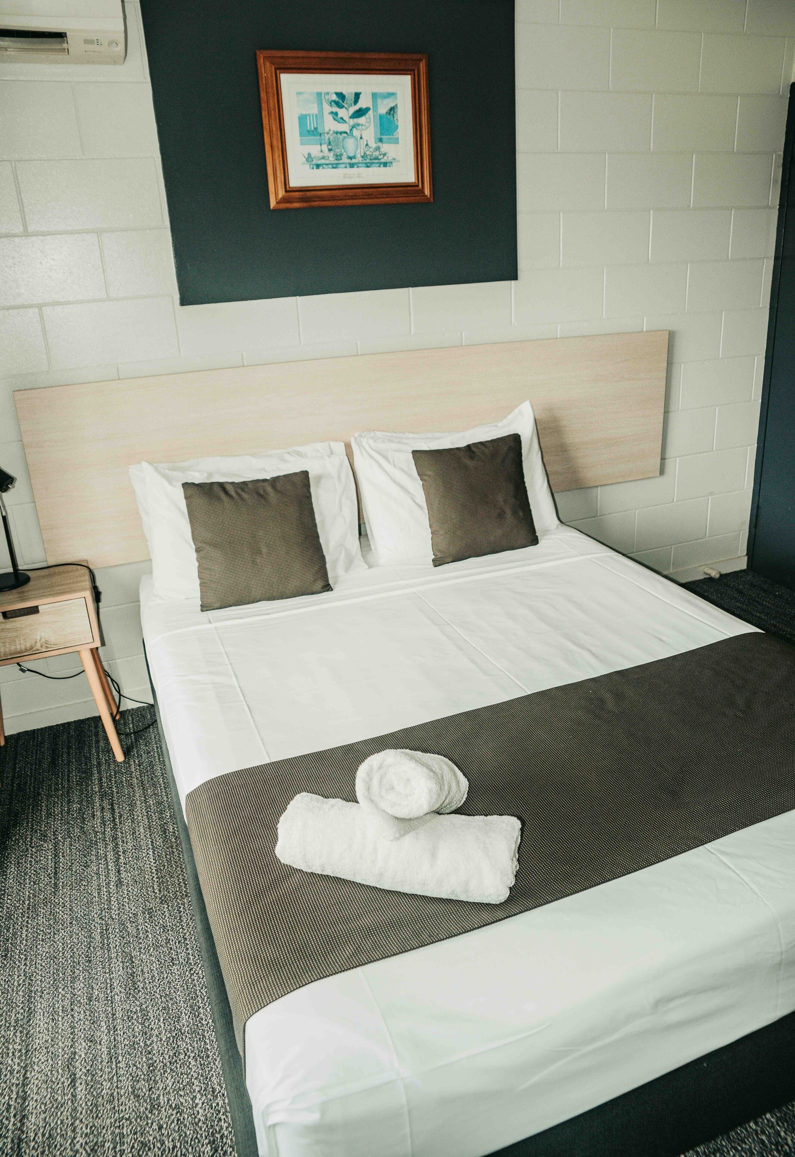 Una cama bien hecha con sábanas blancas y dos almohadas marrones, un cuadro en la pared y una mesita de noche con una lámpara.