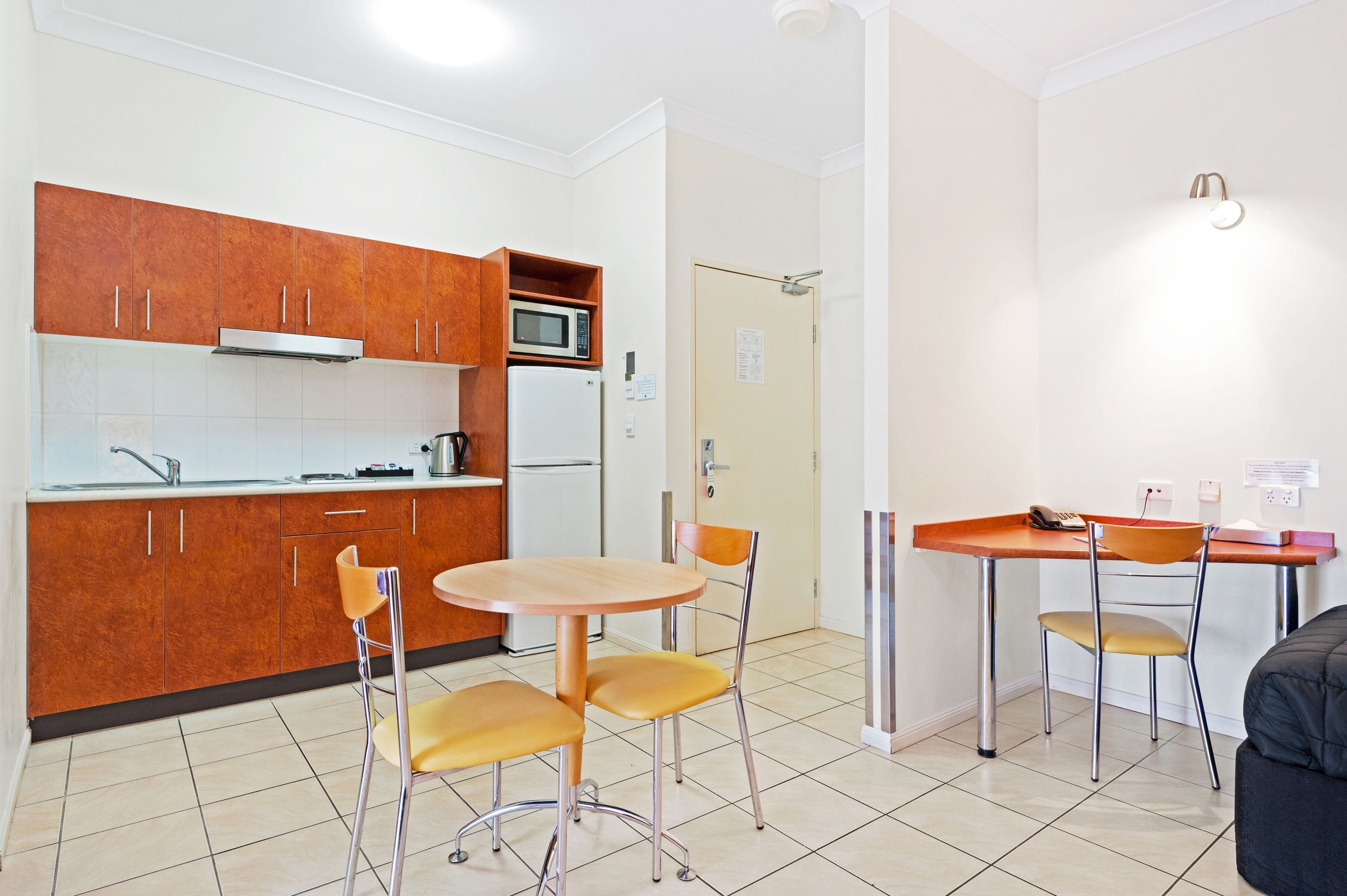Una cocina compacta con armarios de madera, microondas y refrigerador. Cuenta con un pequeño comedor con una mesa y dos sillas.