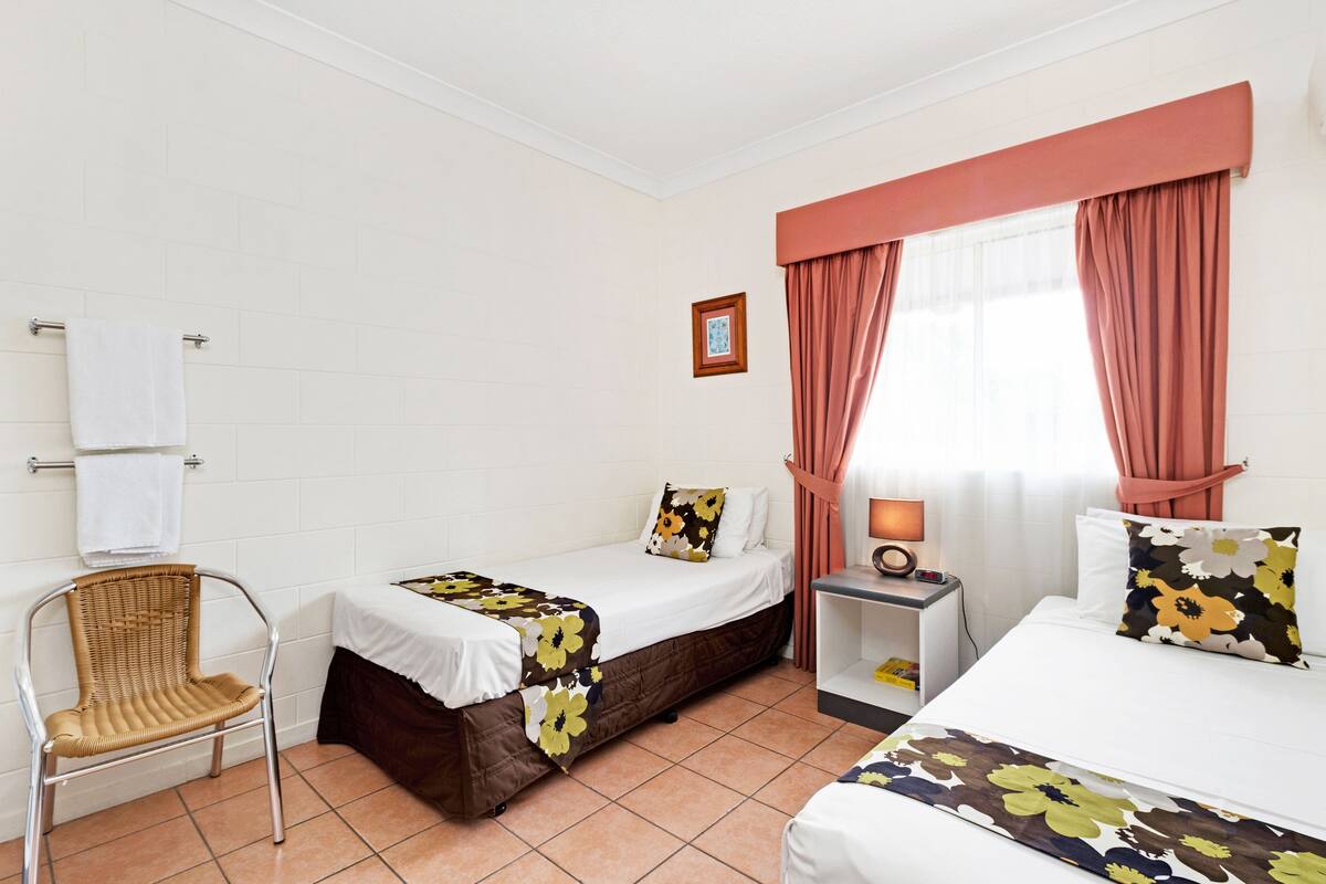 Une chambre d’hôtel avec deux lits, une chaise, une table de chevet et une fenêtre avec des rideaux.